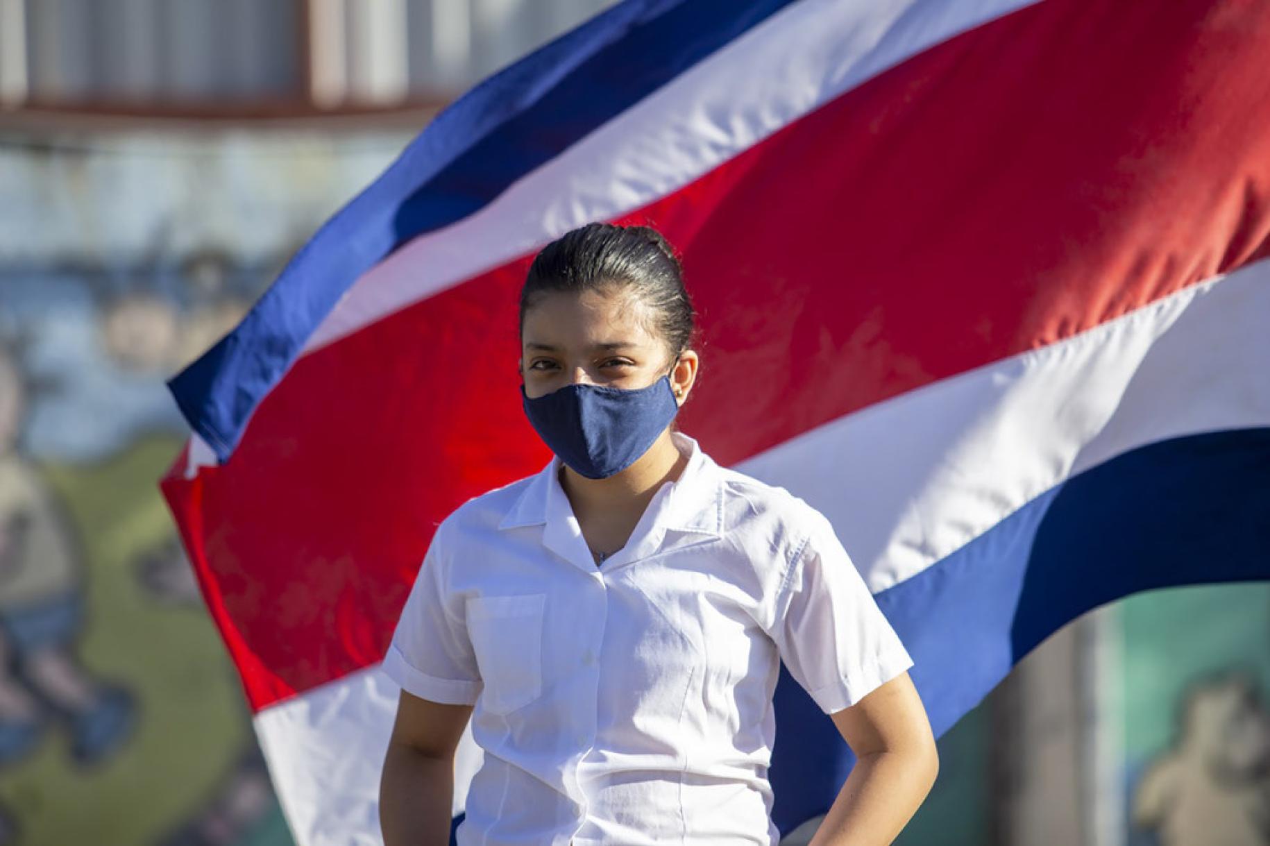 Una niña con una mascarilla azul marino se encuentra parada frente a la bandera de Costa Rica.