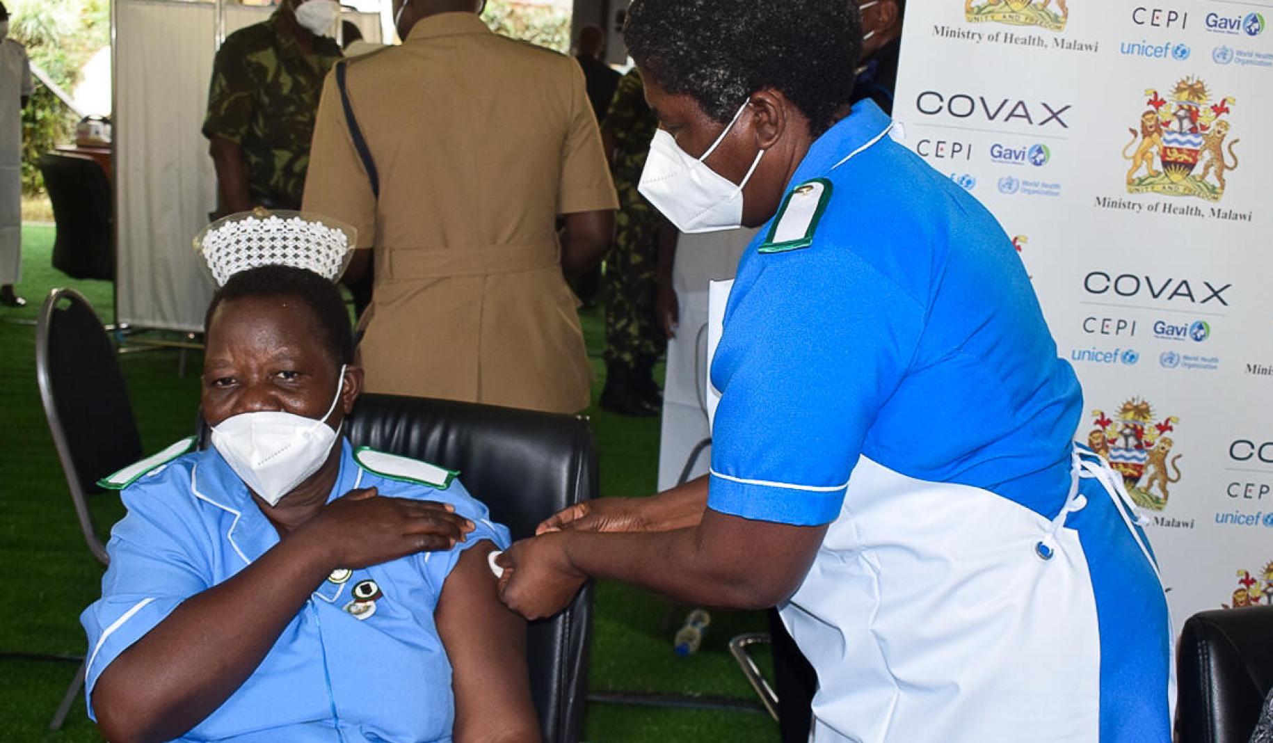 Una profesional de la salud en una locación de vacunación se arremanga el uniforme mientras recibe una vacuna, administrada por su compañera de trabajo.