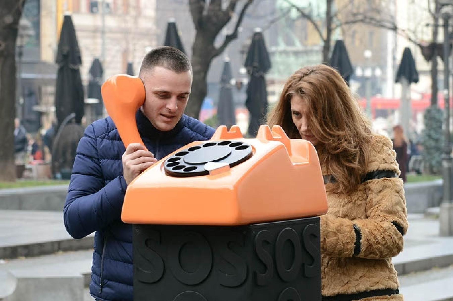 Un hombre y una mujer se sitúan junto a un gran teléfono naranja de discar. El hombre sostiene el auricular. 