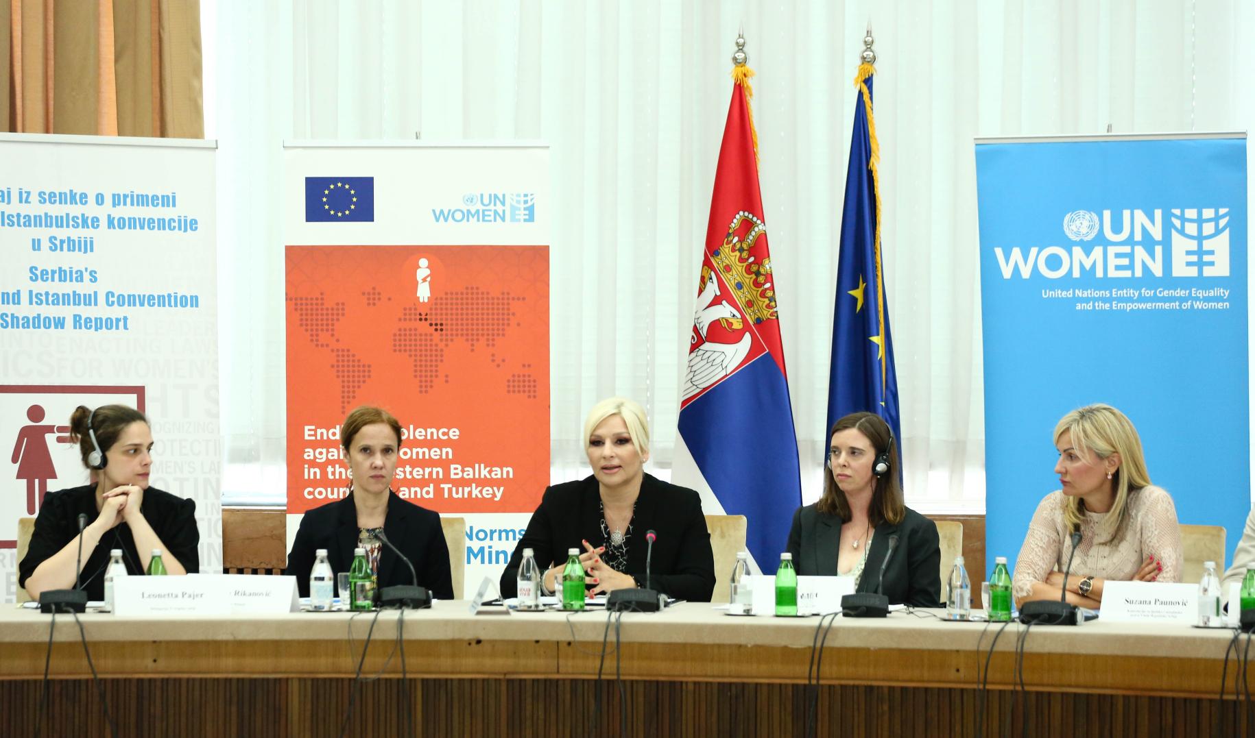Cinco mujeres celebran una conferencia de prensa frente a la bandera de ONU-Mujeres y las banderas.