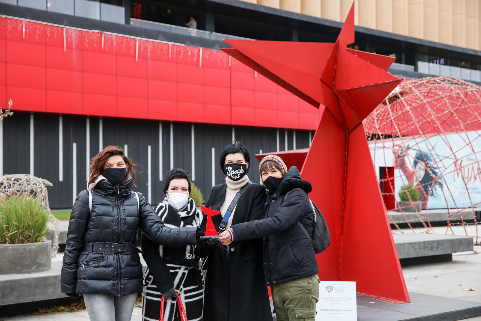 Four women stand together with masks in front of a red statute. 