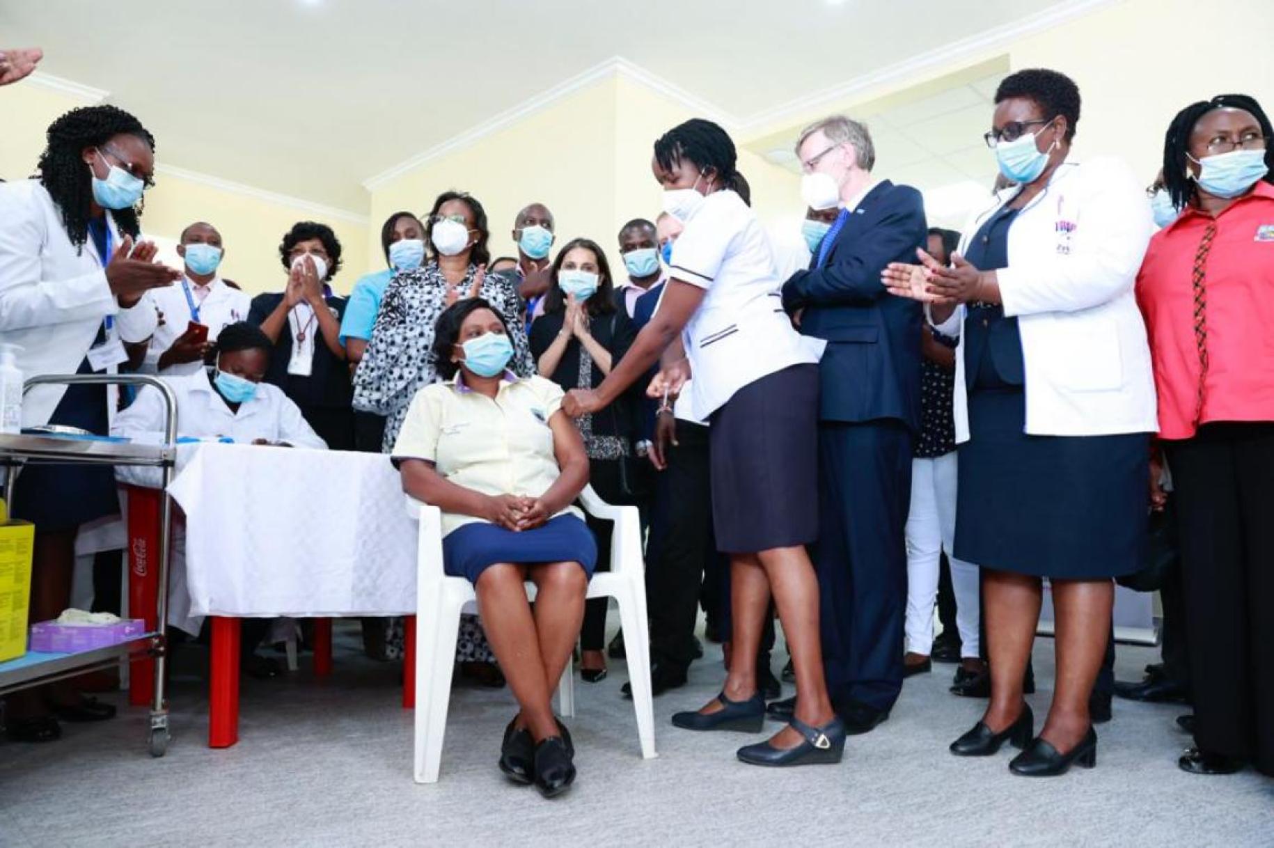 A large group of people wearing masks stand around a seated woman getting vaccinated. 