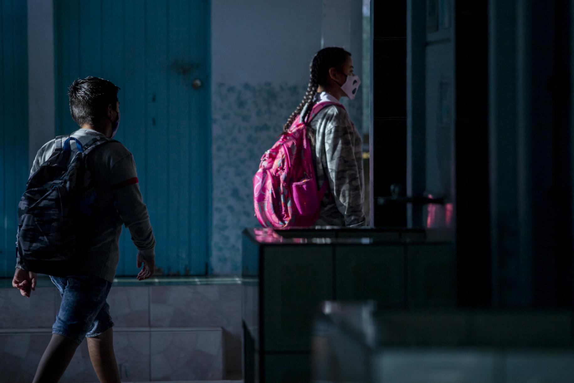 Una niña y un niño entran a la escuela con mochilas y mascarillas.