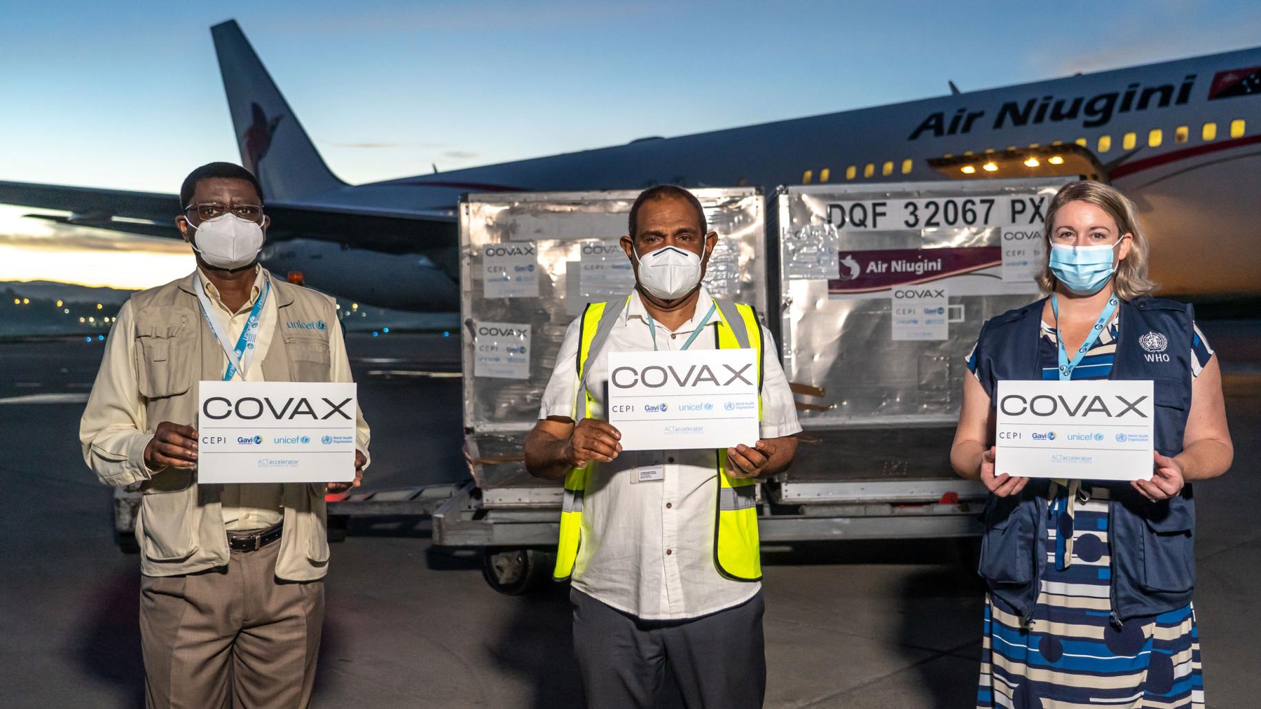 Deux hommes et une femme portant des masques de protection respiratoire et tenant des panneaux de l'initiative COVAX se tiennent debout, face caméra, sur le tarmac d'un aéroport où vient d'atterrir un avion chargé de vaccins anti-COVID-19.