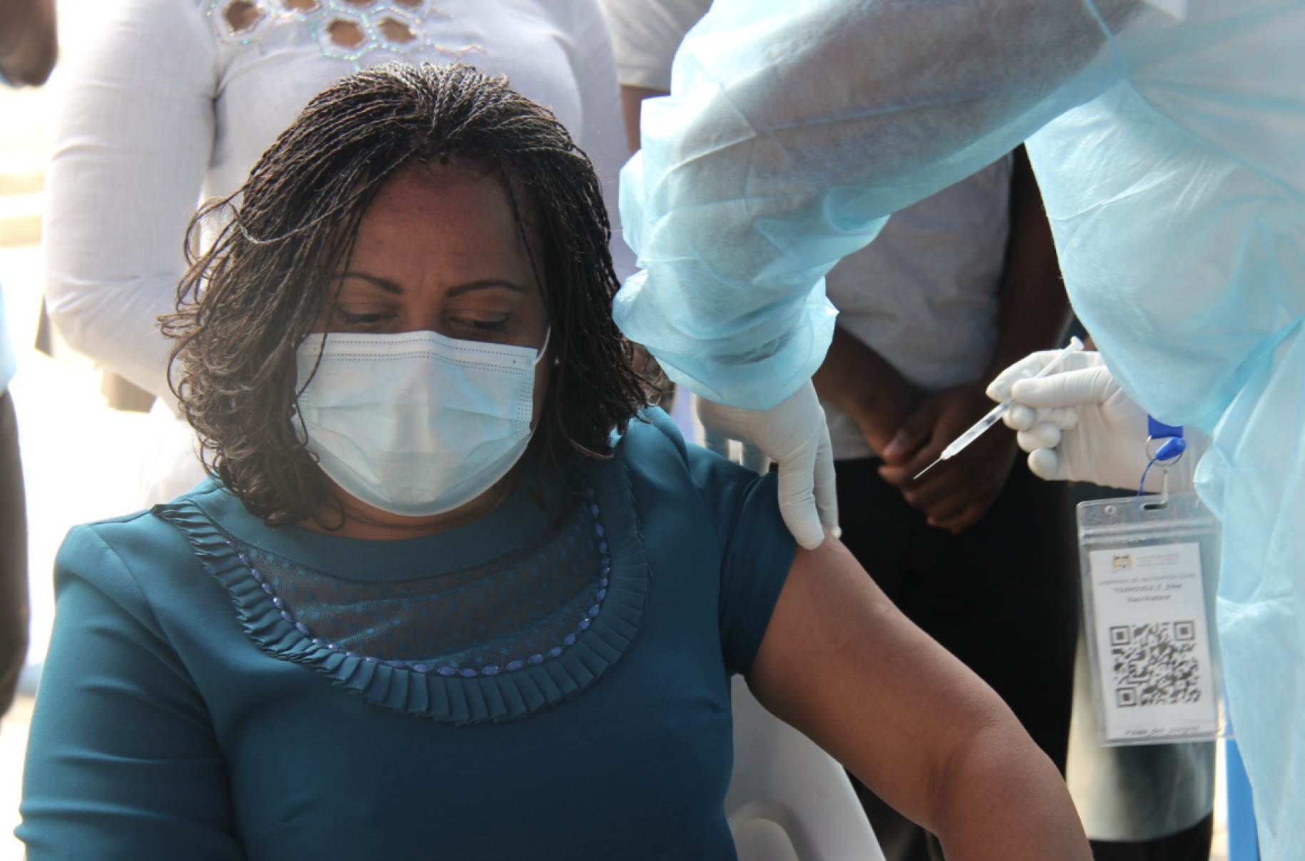 A woman wearing a a teal shirt, looks down while she is vaccinated by a person in protective equipment. 