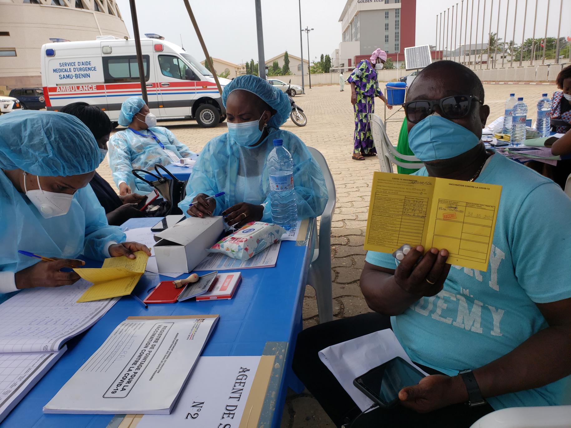 A man in sunglasses, a blue shirt, and a blue face mask shows his vaccination form to the camera while two health care workers sit near him.