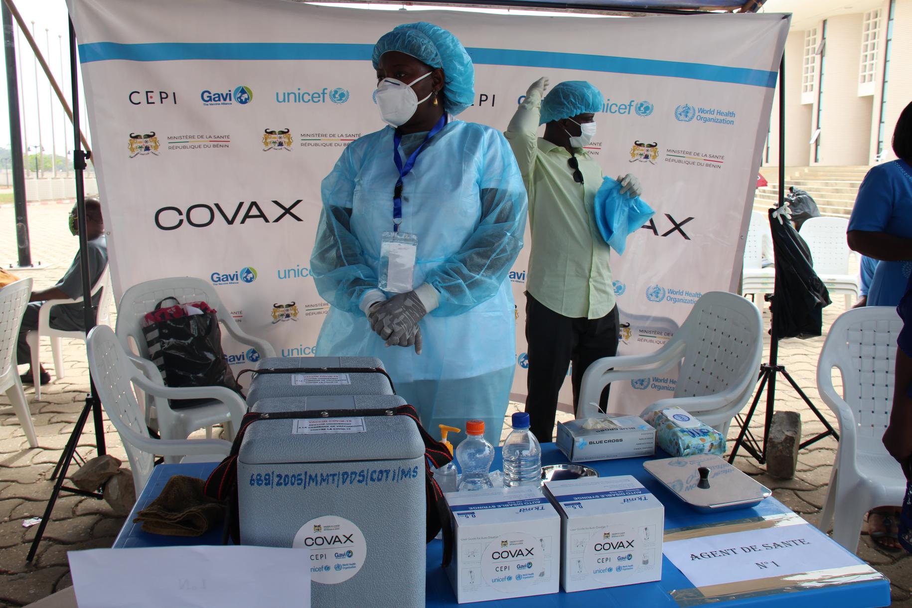 Two people in protective medical gear stand near vaccination supplies. 