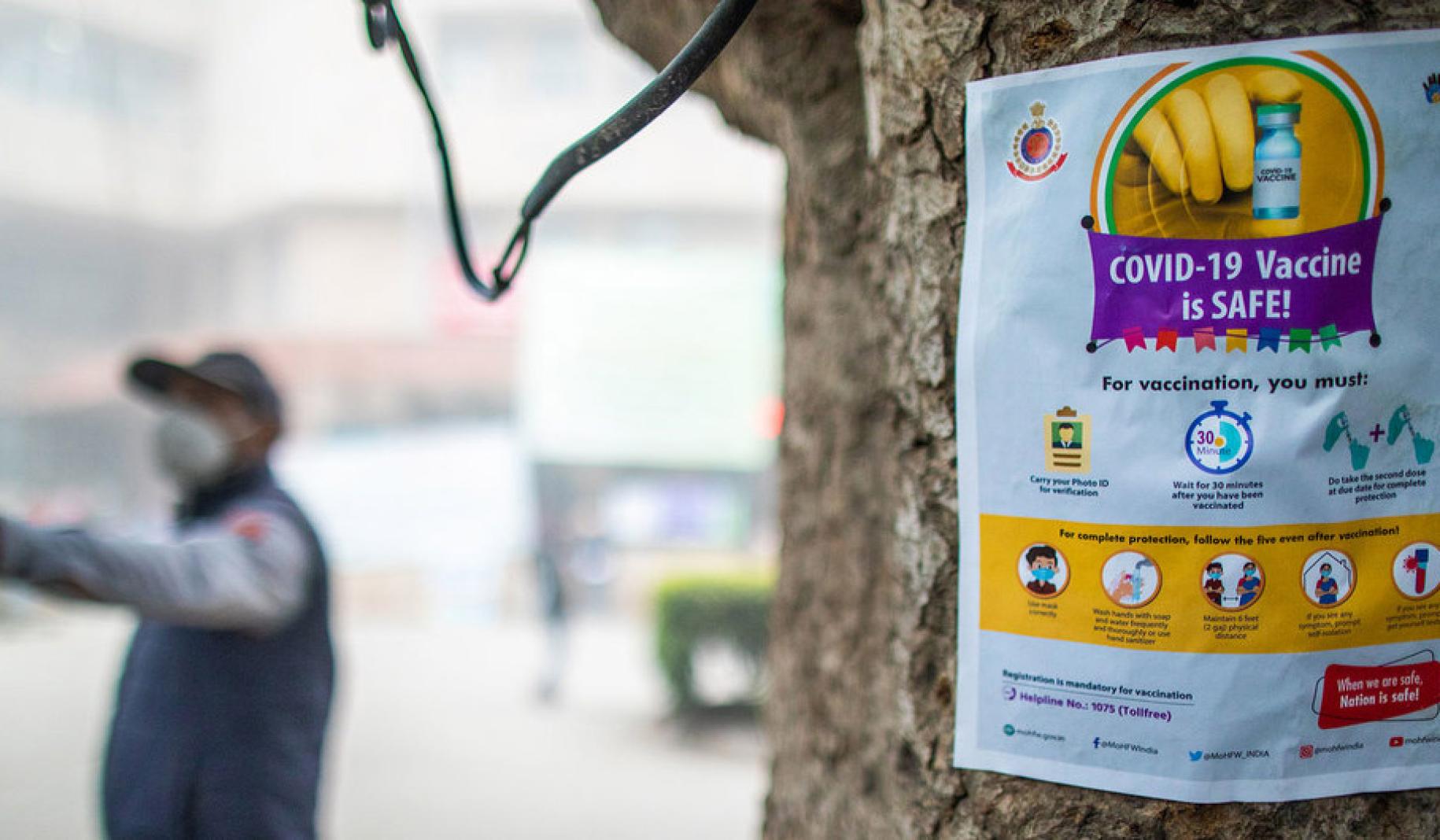A sign, with the image of a vaccine and information, is posted on a tree and a person in a mask is seen in the background pointing. 