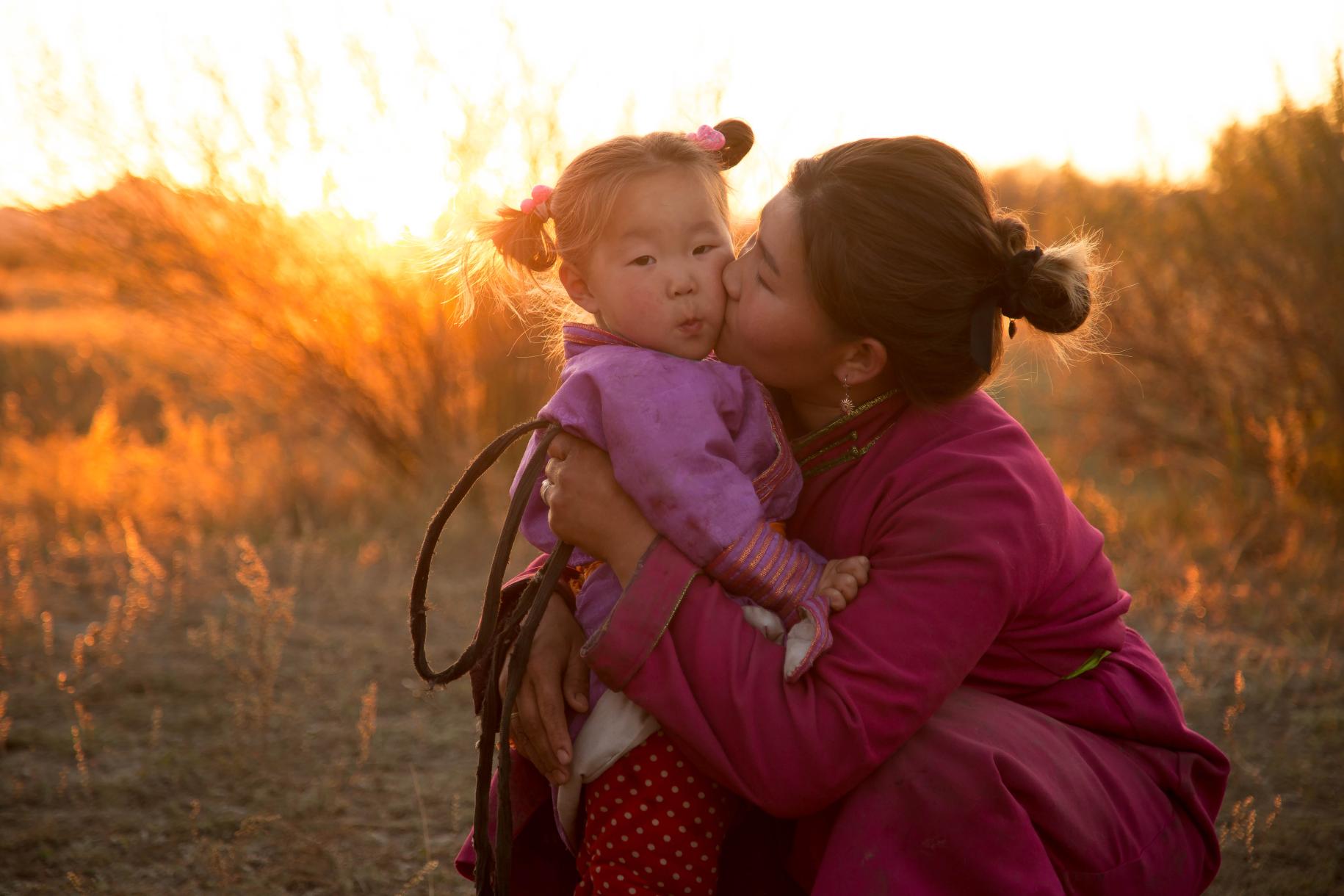 Une femme s'accroupit pour embrasser une petite fille sur la joue dans un champ, au moment du coucher du soleil.