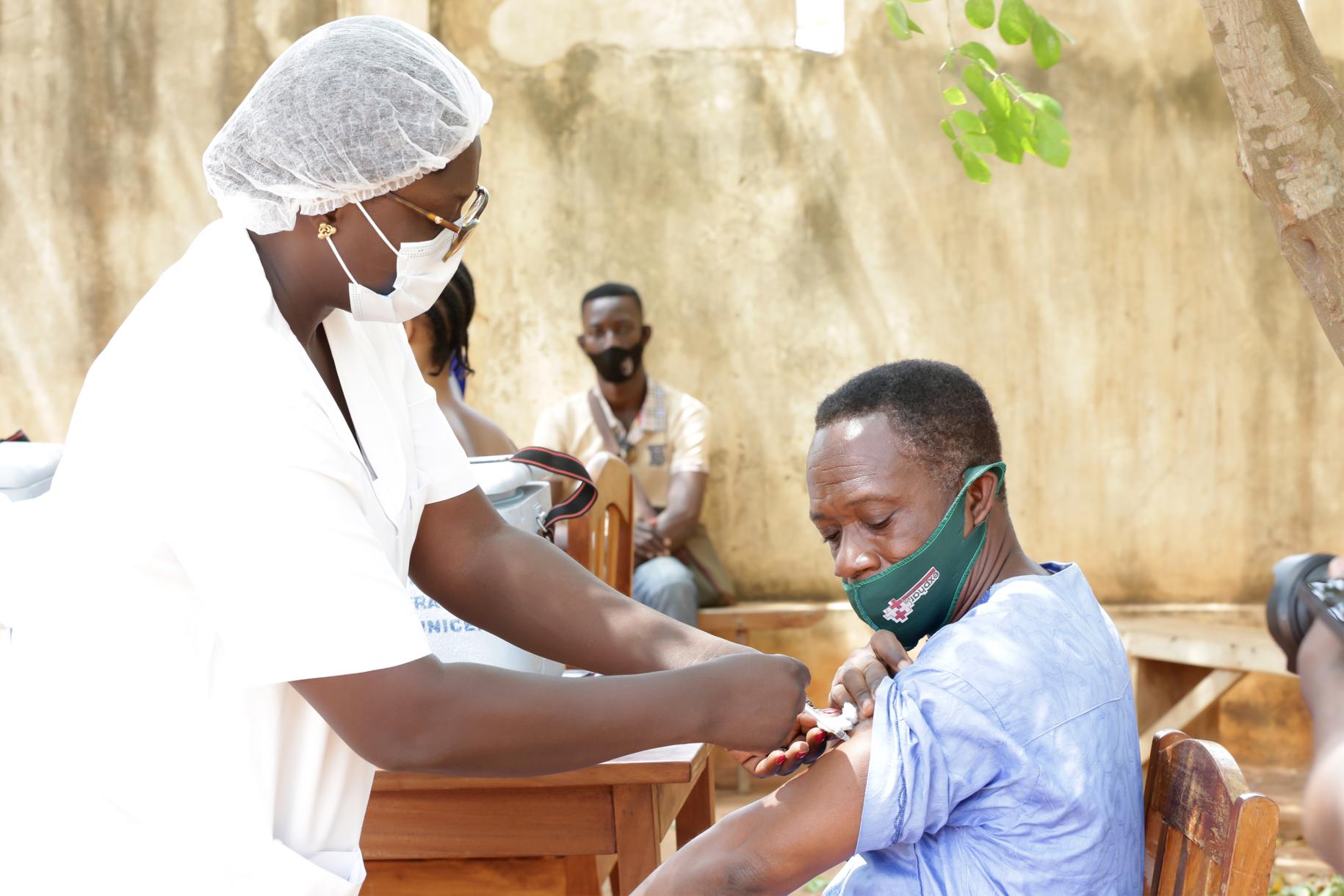 A man with a green mask receives a Covid-19 vaccine from a woman dressed in medical gear. 