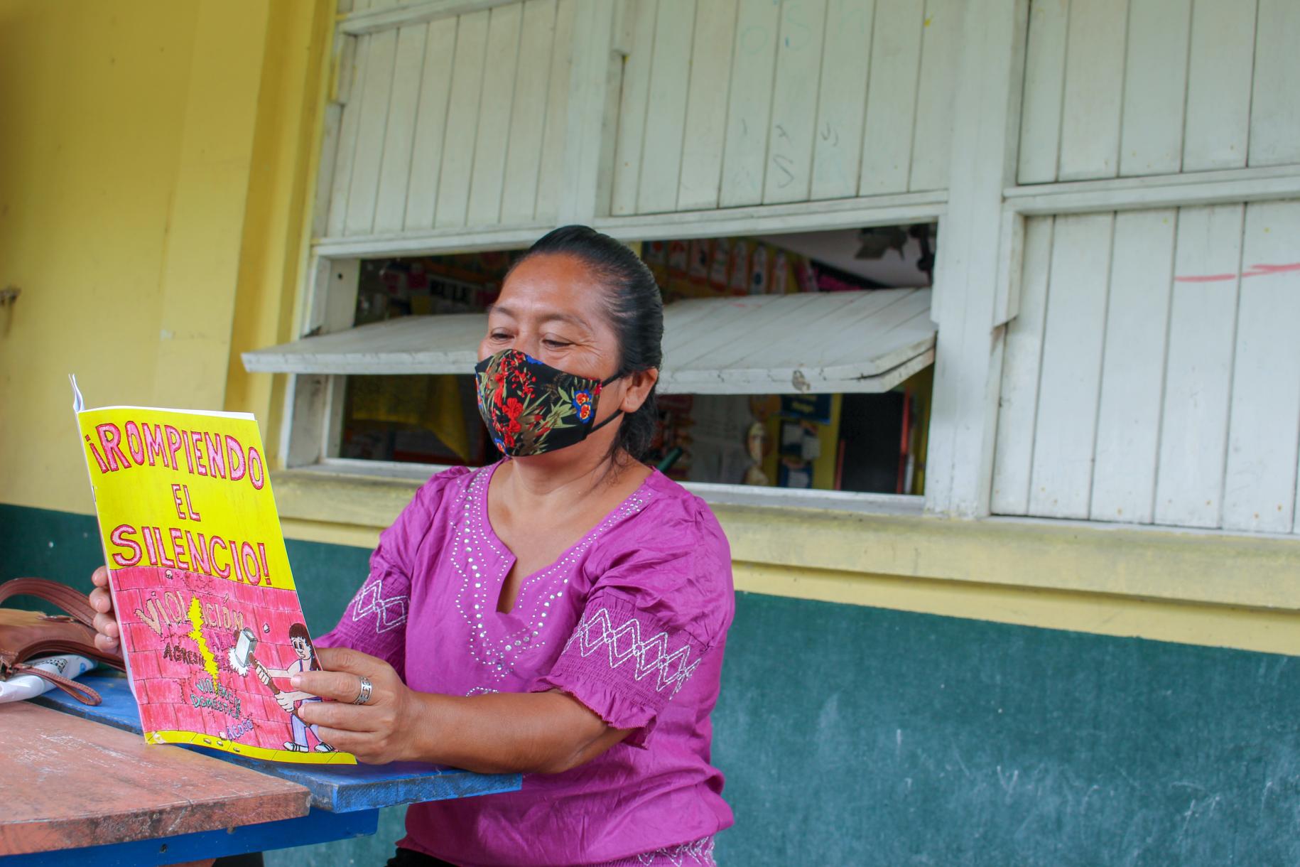 A woman in a colorful face mask and purple shirt reads a book while sitting at a table.