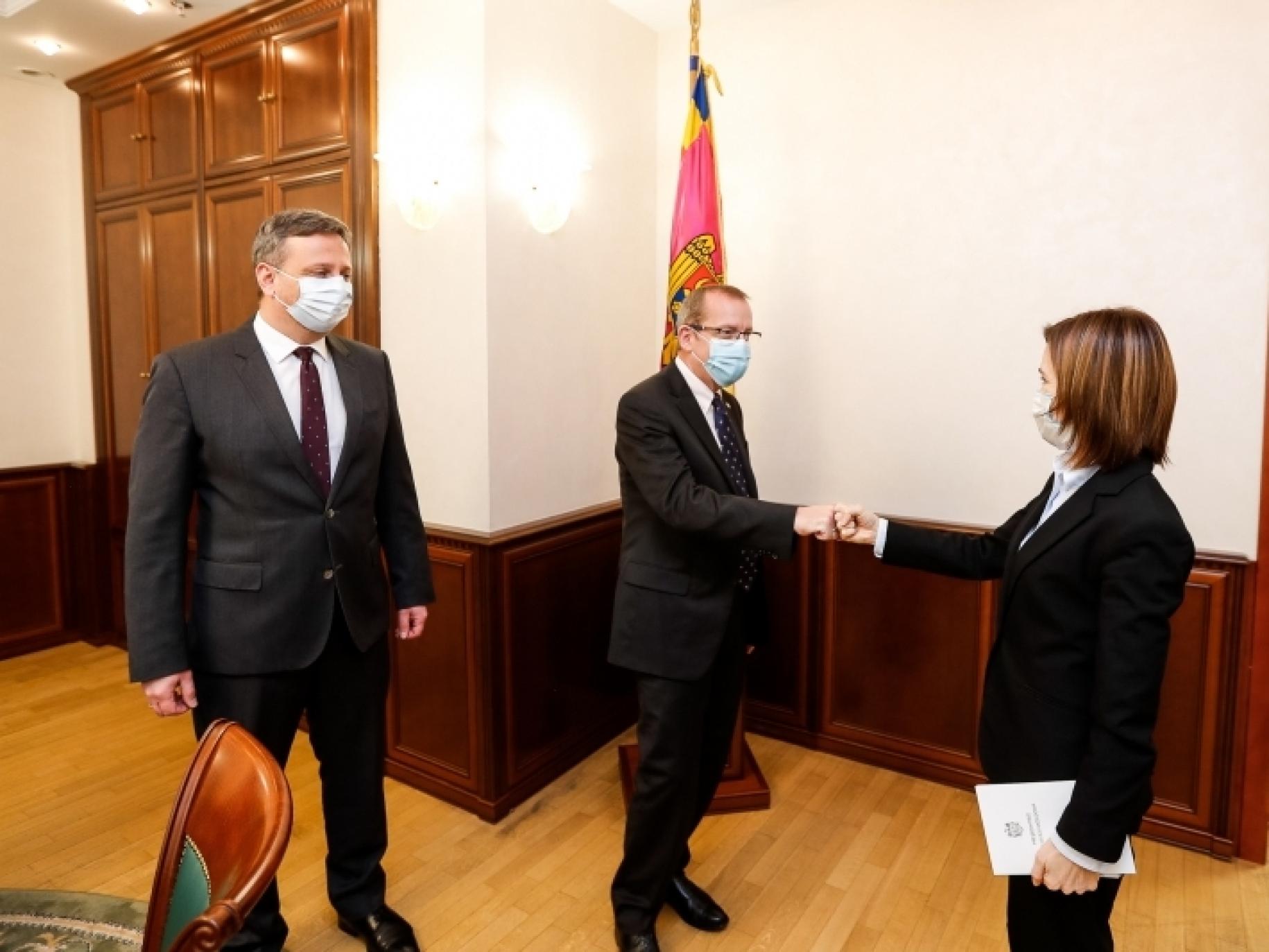 Un homme en costume et une femme en tailleur se serrent la main tandis qu'un autre homme se tient debout à côté d'eux. Tous trois portent un masque de protection respiratoire.