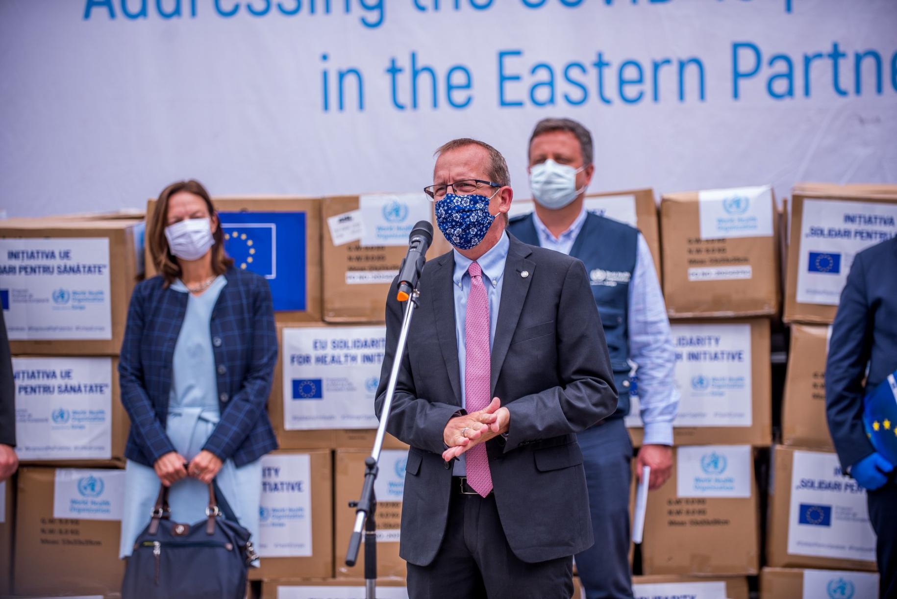 Four people in face masks stand in front of COVAX Vaccine boxes while one man speaks at a microphone. 