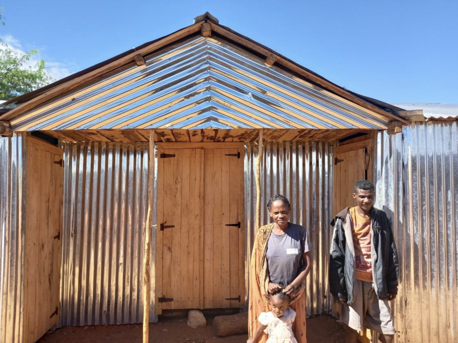 The image shows Fagnosea and Masy with their child standing in front of the house they built.