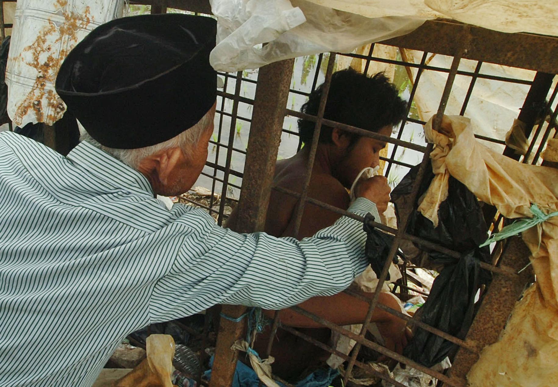 Un joven en una jaula es ayudado por un hombre mayor con sombrero negro fuera de la jaula. 