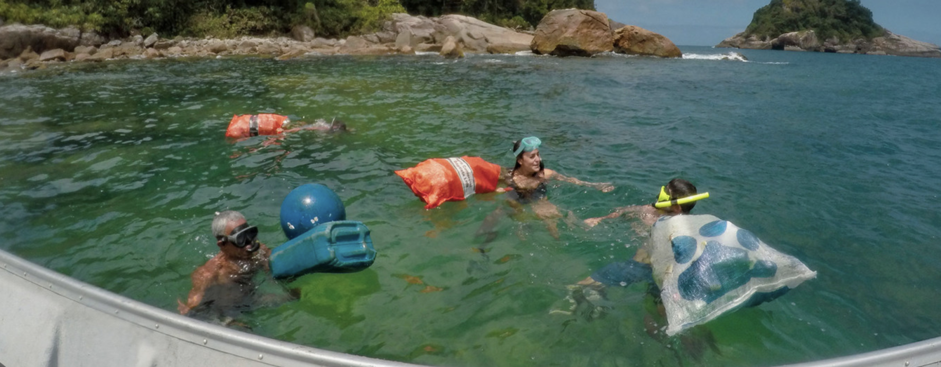 Several people swim in the ocean with large bags for waste collection and a floating plastic buoy.