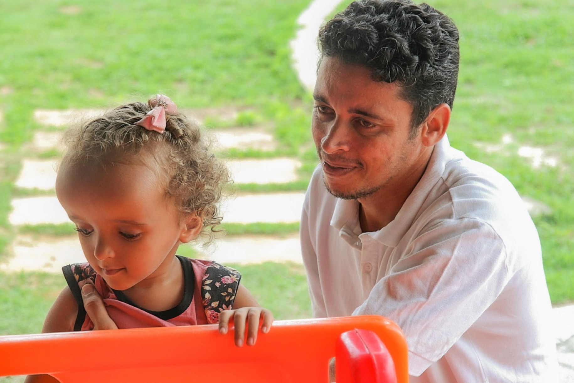 Un hombre y una niña pequeña observan juntos en dirección a un juguete naranja, en un espacio exterior con jardín.