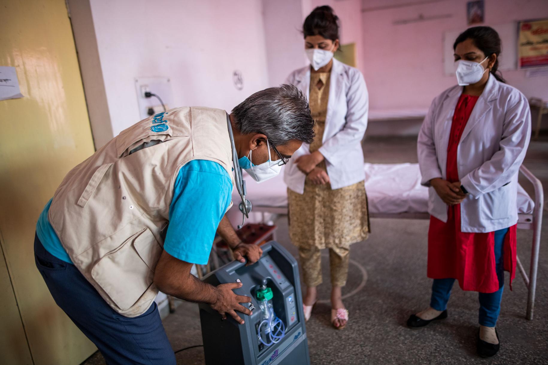 Un hombre, con un chaleco de UNICEF, con un concentrador de oxígeno muestra a dos profesionales médicos cómo funciona.