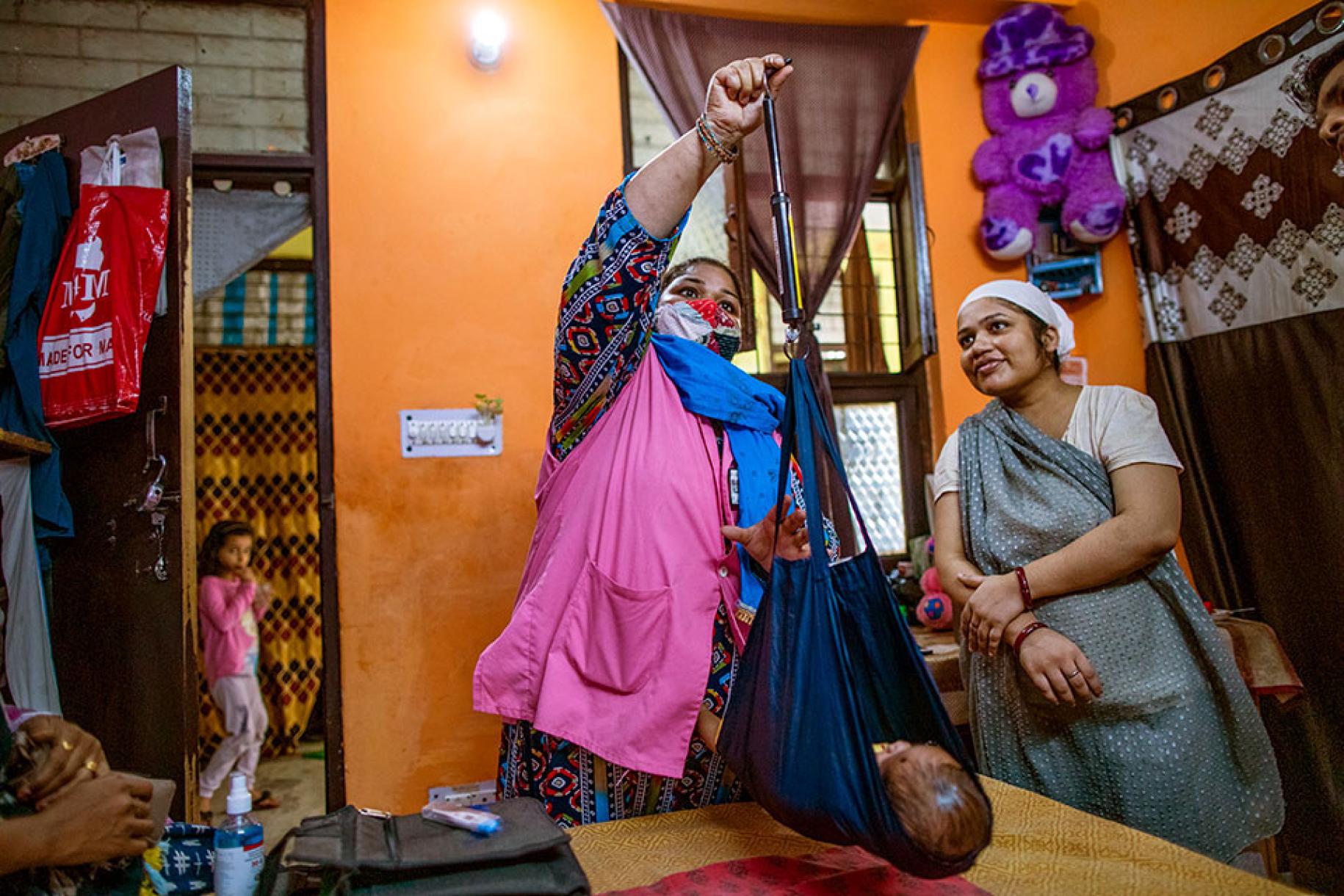 Une agente de santé accréditée pèse un bébé dans le cadre d’une des visites à domicile qu’elle effectue pour faire des bilans de santé pré et postnataux à New Delhi, en Inde. La mère de l’enfant se tient debout, près d’elle, et la regarde faire en souriant.