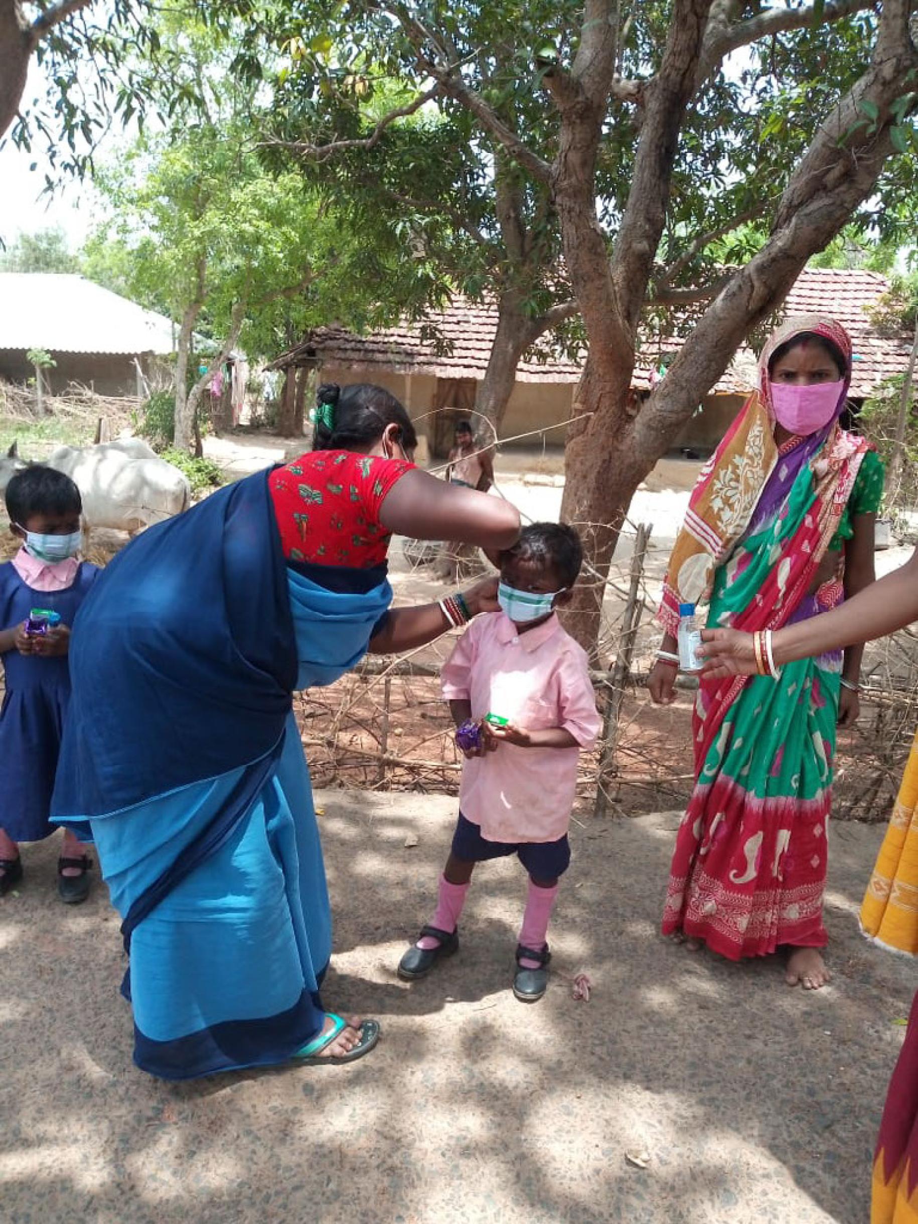 En Inde, à l’ombre d’un arbre ; une femme portant un sari aide une petite fille à ajuster son masque de protection respiratoire, tandis qu’une autre femme, en sari elle aussi, se tient debout face caméra et qu’une troisième, hors champ, tend un façon de gel hydroalcoolique à la fillette.
