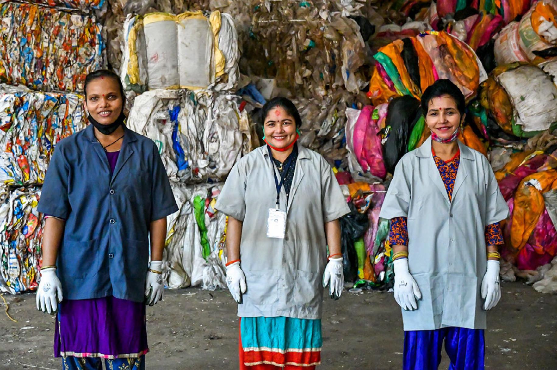 Three women smile at the camera in front of large piles of packaged waste.