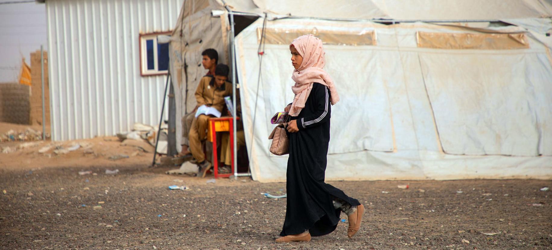 Une jeune fille yéménite vêtue de noir et portant un voile de couleur rose clair sur la tête se déplace dans un camp de réfugiés et passe devant une tente de couleur beige.