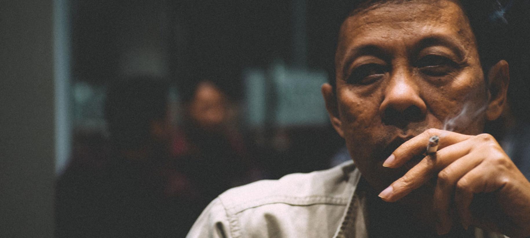 Gros plan sur le visage d'un homme d'origine asiatique en train de fumer une cigarette dans une pièce sombre.