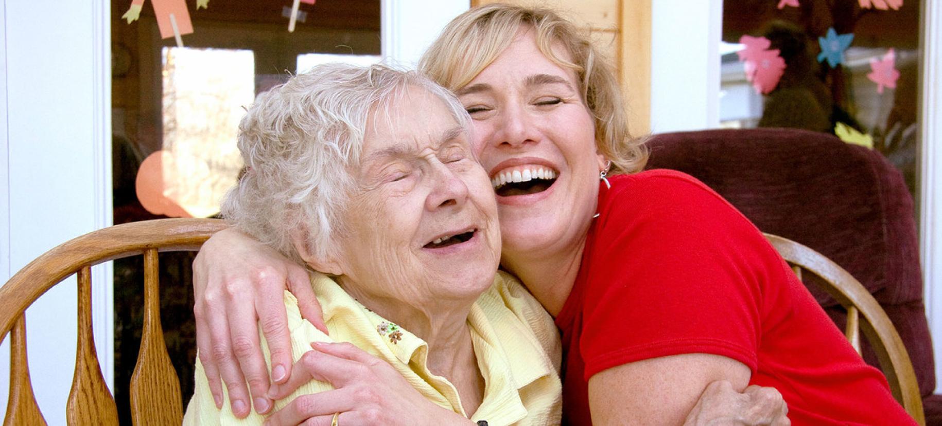 Une jeune femme blonde en t-shirt rouge et une femme âgée en polo jaune se prennent tendrement dans les bras l'une de l'autre, avec un grand sourire.