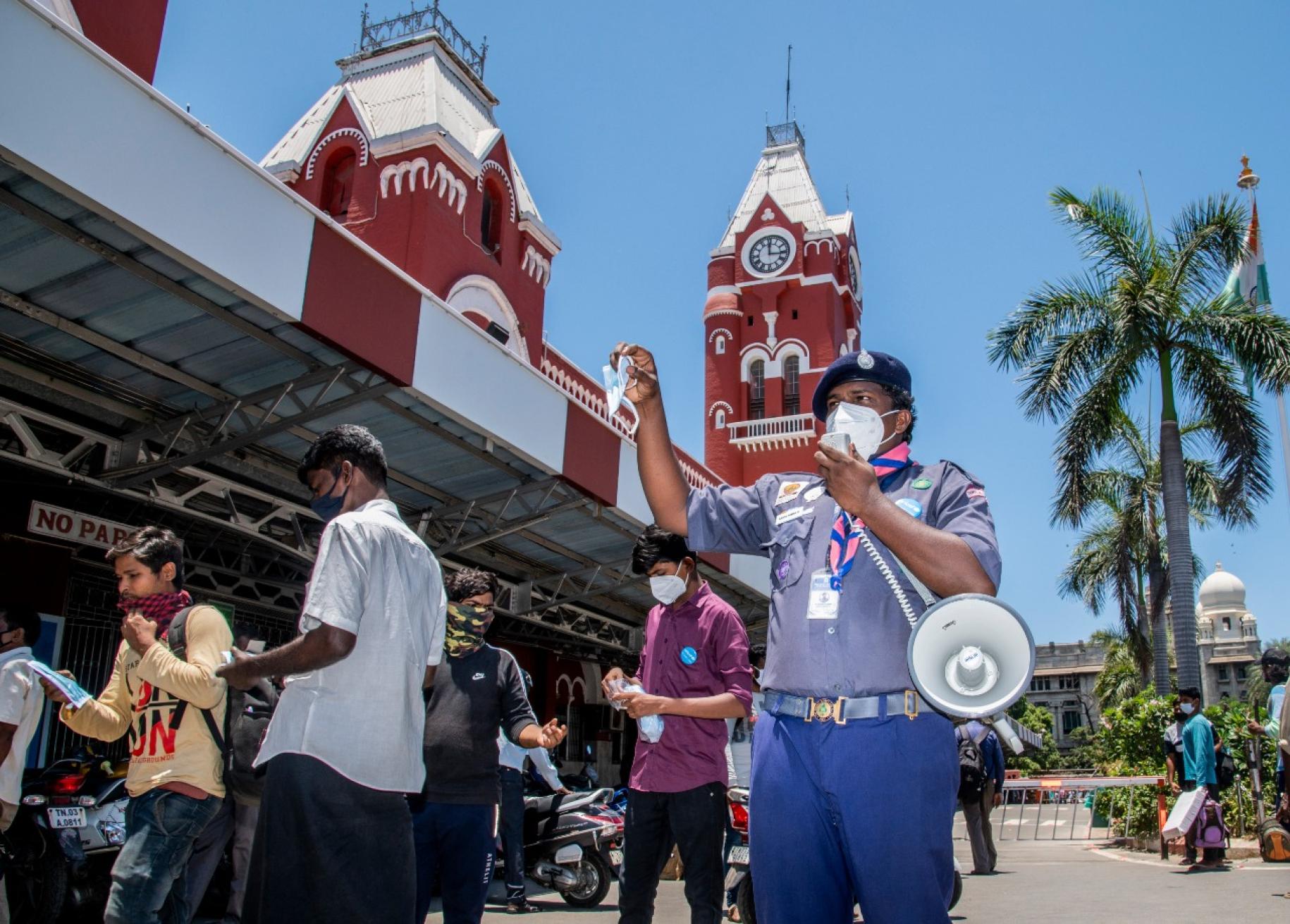 Un homme en uniforme muni d'un mégaphone passe devant un beau bâtiment rouge, dans une rue bordée de palmiers, en Inde, pour faire passer aux habitants des messages sanitaires sur les gestes barrières, les traitements et les raisons d’avoir confiance en la vaccination contre la COVID-19.