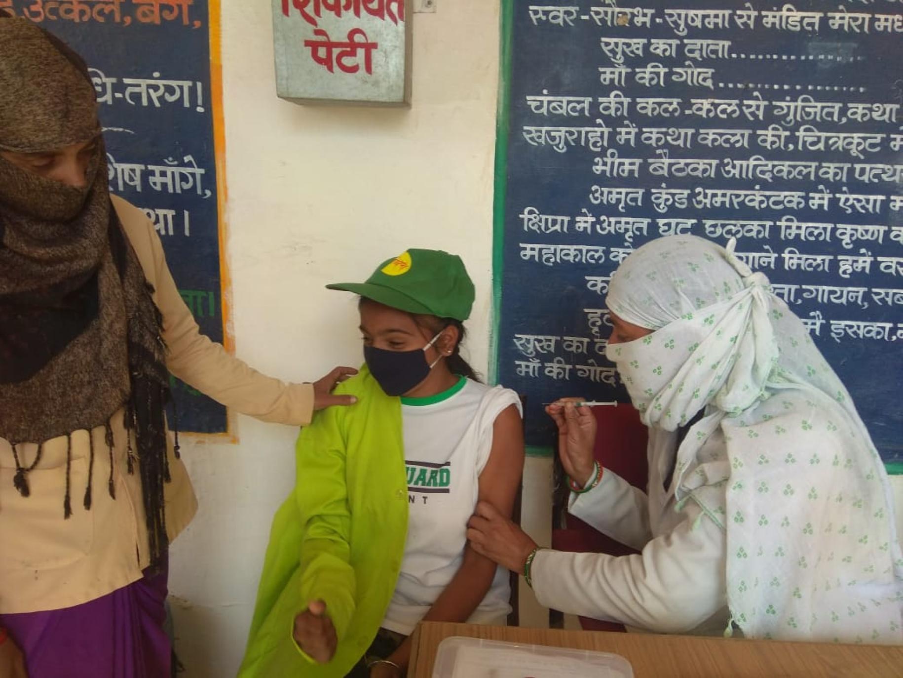 Dans une école du district de Chattarpur, à Madhya Pradesh, en Inde, une jeune fille portant une casquette verte et un masque de protection respiratoire se fait vacciner contre la COVID-19 par une infirmière voilée, alors qu’une autre femme, voilée également, pose la main sur l’épaule de la jeune fille pour la rassurer. 