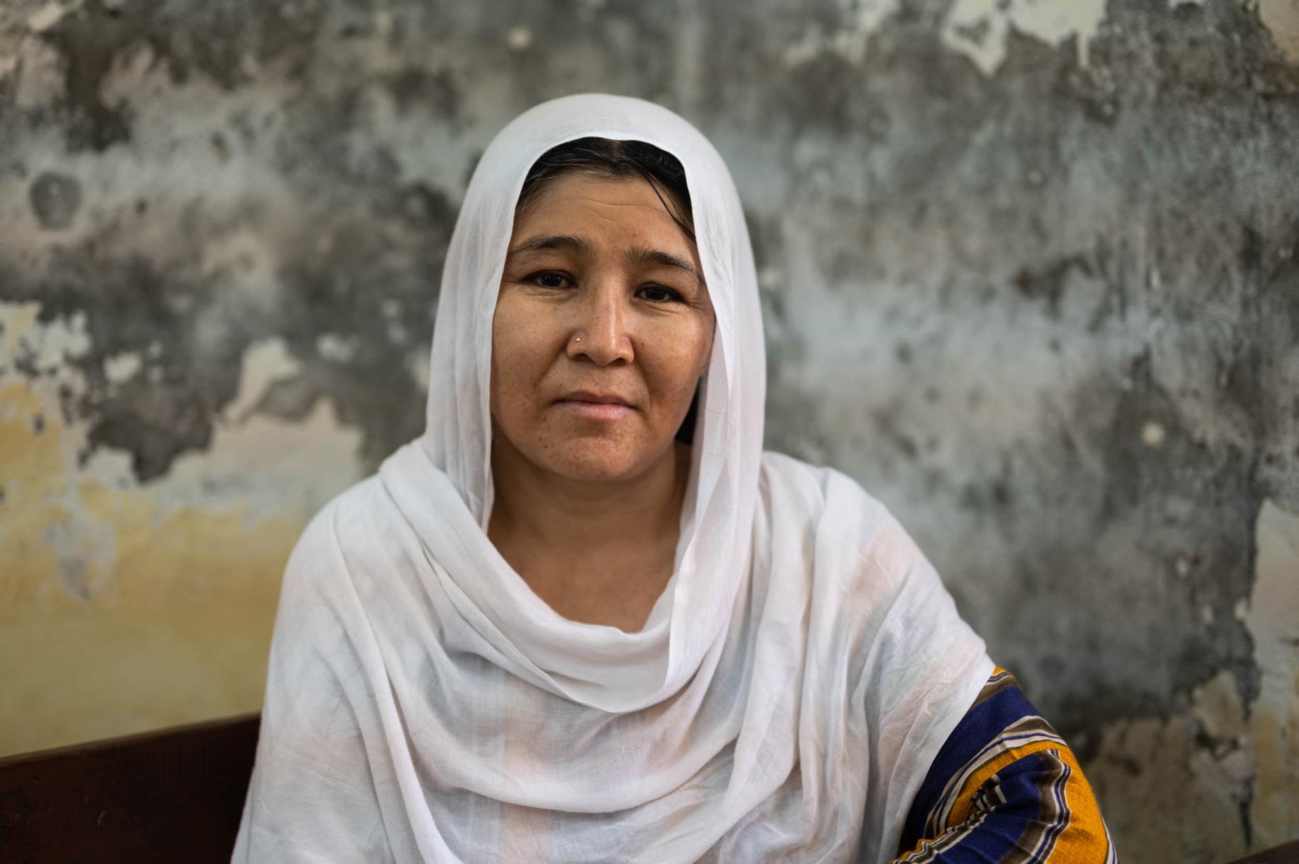 Portrait d’une réfugiée afghane portant un foulard blanc sur la tête et les épaules et assise devant un mur décrépit.