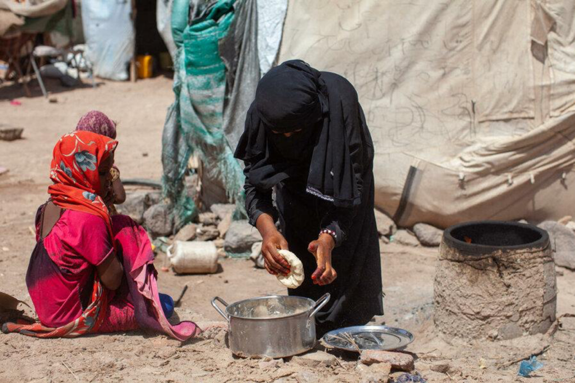 Une femme portant une longue robe noire et un voile noir sur la tête se penche en avant sur une marmite posée au sol, devant une tente, dans un camp de réfugiés, au Yémen.