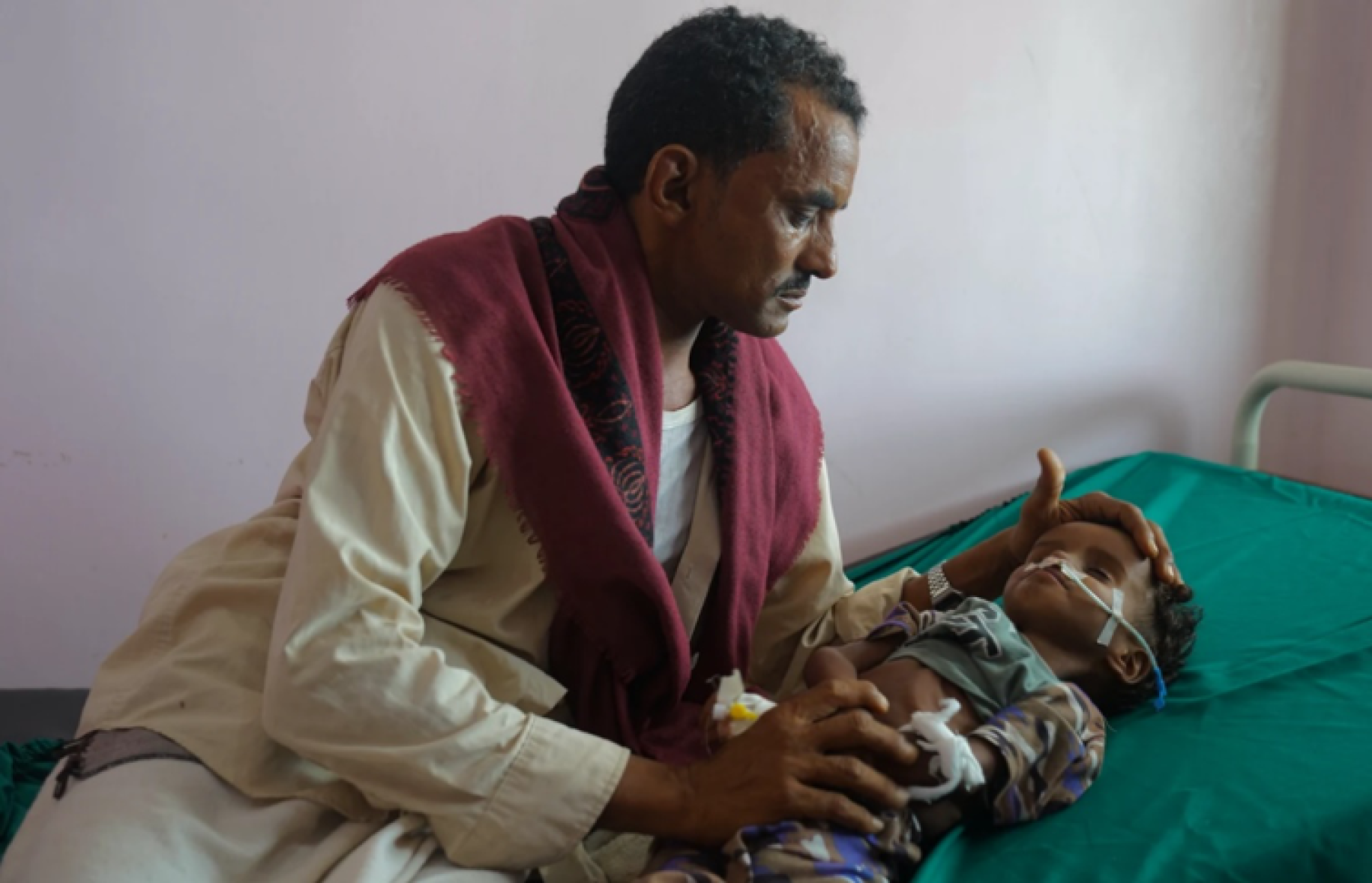 Une jeune enfant souffrant de malnutrition aiguë est allongé sur un lit d’hôpital tandis que son oncle, le visage fermé, se penche sur lui et pose les mains sur sa tête et ses pieds.