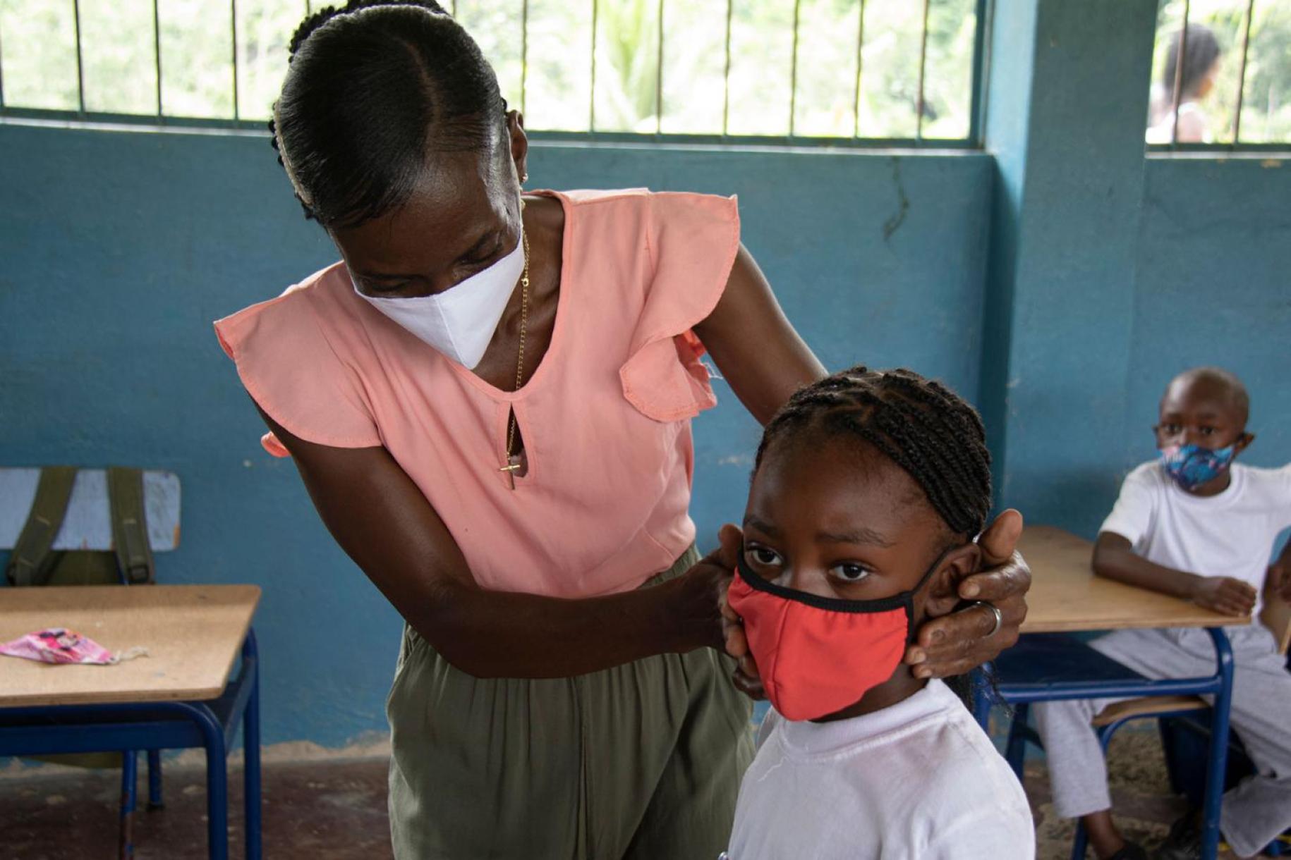 Gros plan sur une enseignante de l’enseignement primaire portant un masque de protection respiratoire qui aide un élève à ajuster son masque sur le visage dans une salle de classe.