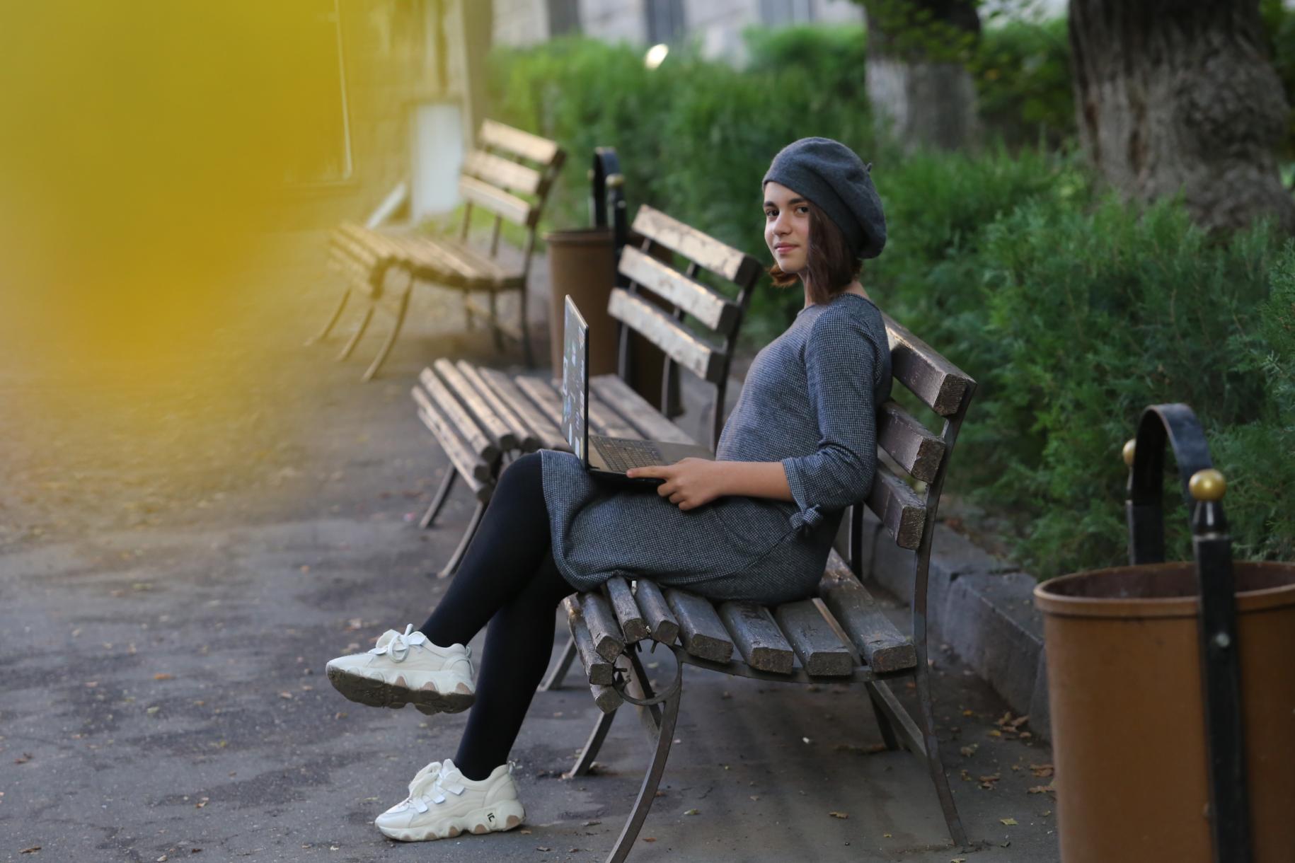 Une jeune fille portant un béret est assise sur un banc, dans un parc, un ordinateur portable sur les genoux, et se tourne vers la caméra en souriant.