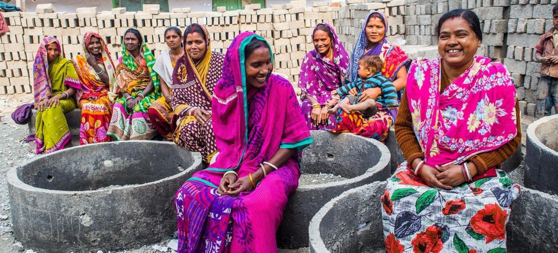 Mujeres vestidas con saris de colores se sientan y hablan, con un fondo de ladrillos para una futura estructura detrás de ellas.