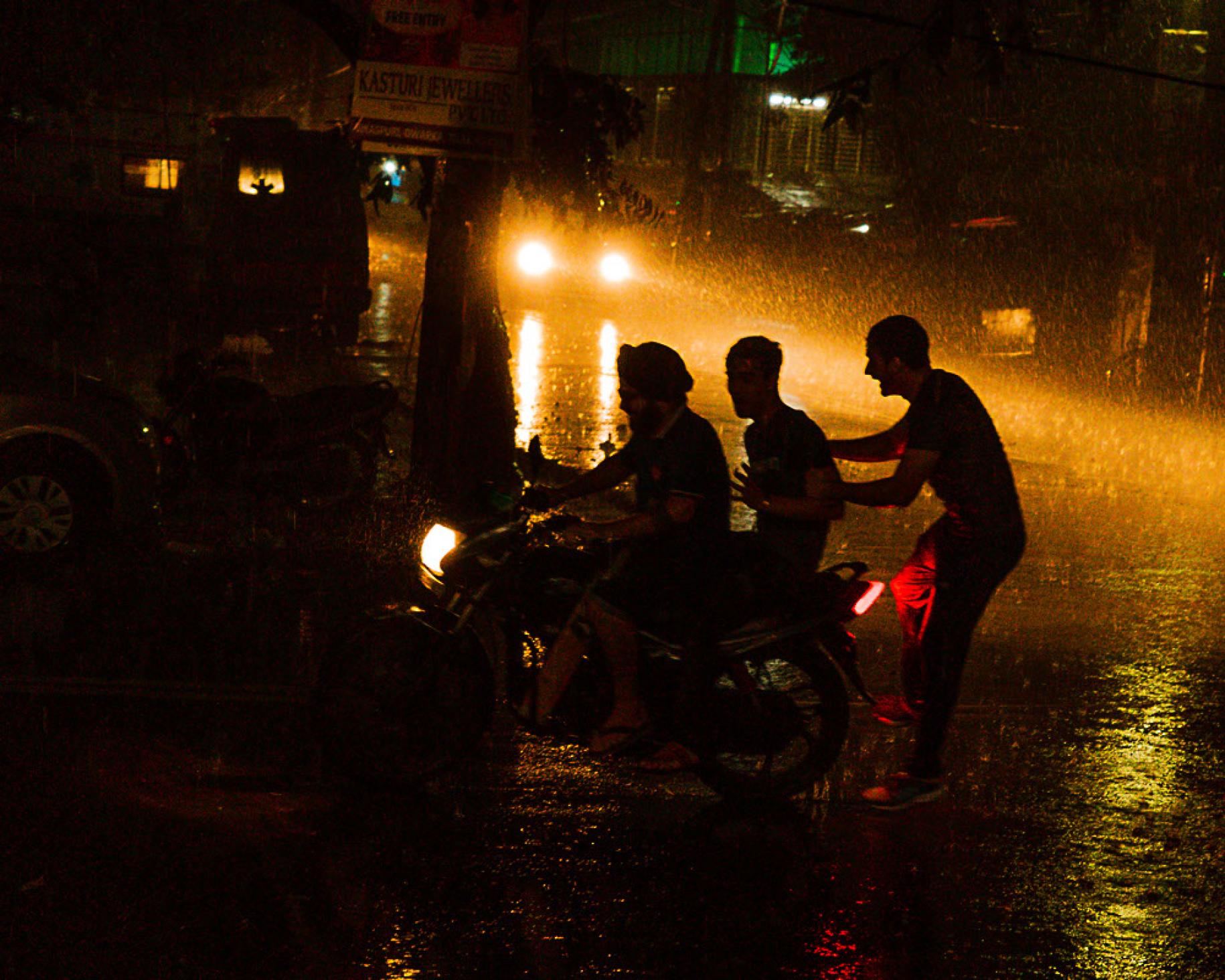 La silueta de tres personas, a contra luz, riendo bajo la lluvia en la calle, durante la noche, con dos focos que aclaran la toma desde lejos.