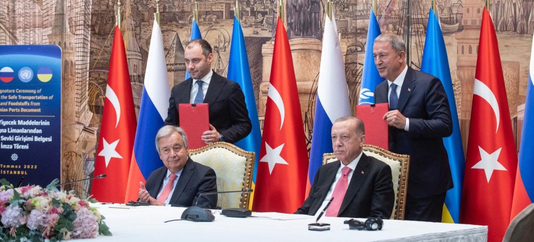 Two men in suits sit at a table with microphones ahead of them, with aides standing behind them. The table is adorned with Russian, Turkish, and UN flags.
