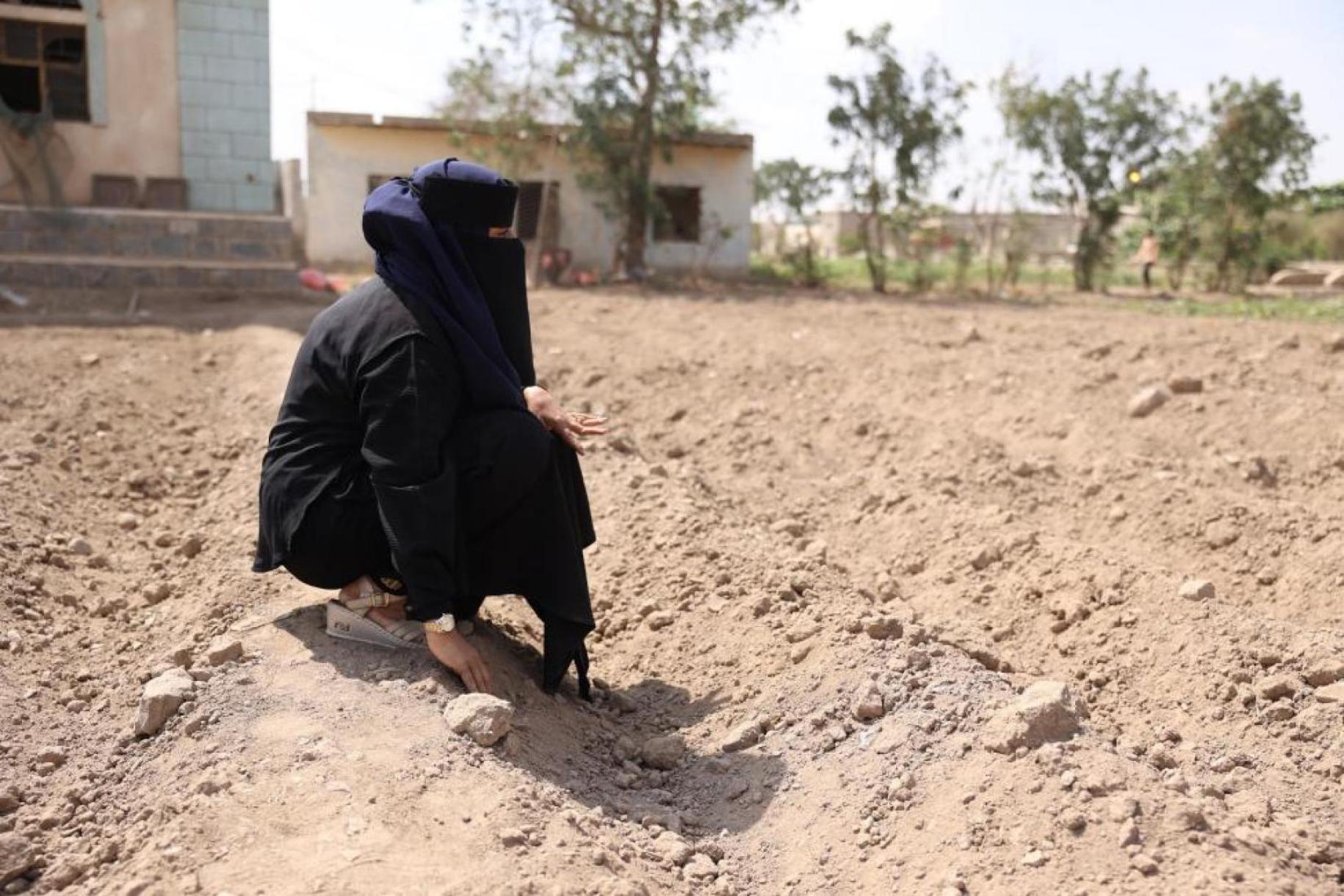 A Lahj, un des gouvernorats du Yémen, une femme entièrement voilée est accroupie au sol sur un terrain extrêmement sec devant des habitations modestes.