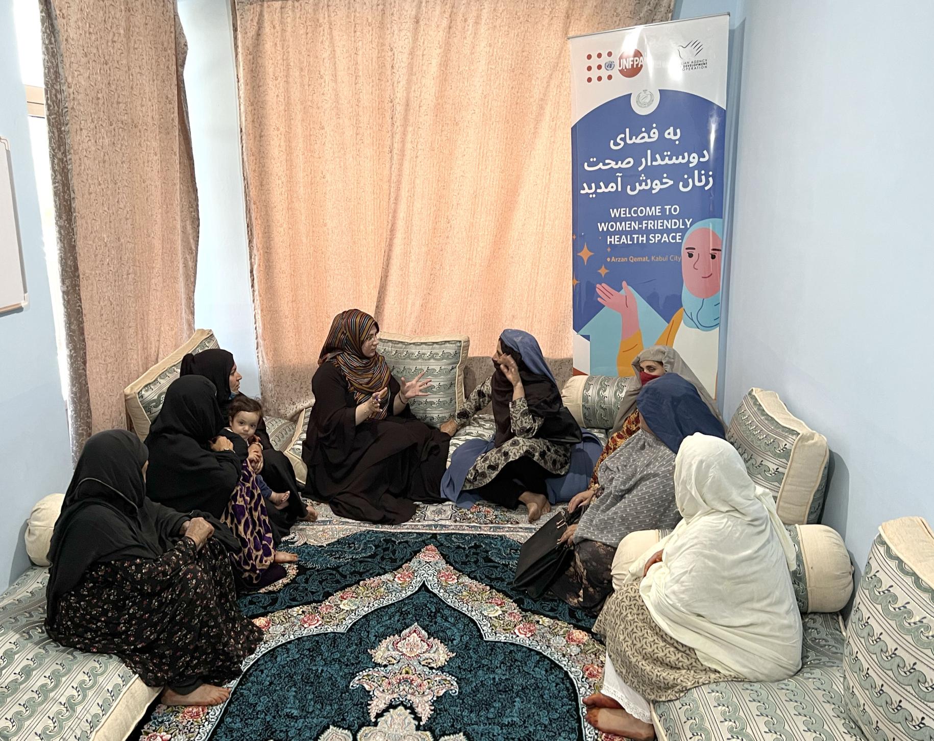 En Afghanistan, un groupe de femmes intégralement voilées sont assises en cercle sur des matelas, à même le sol, dans une pièce fermée, et discutent entre elles. En arrière-plan a été installée la bannière du programme " Welcome to women-friendly health space", qui signifie en français "Bienvenue dans l'espace de santé adapté aux femmes".