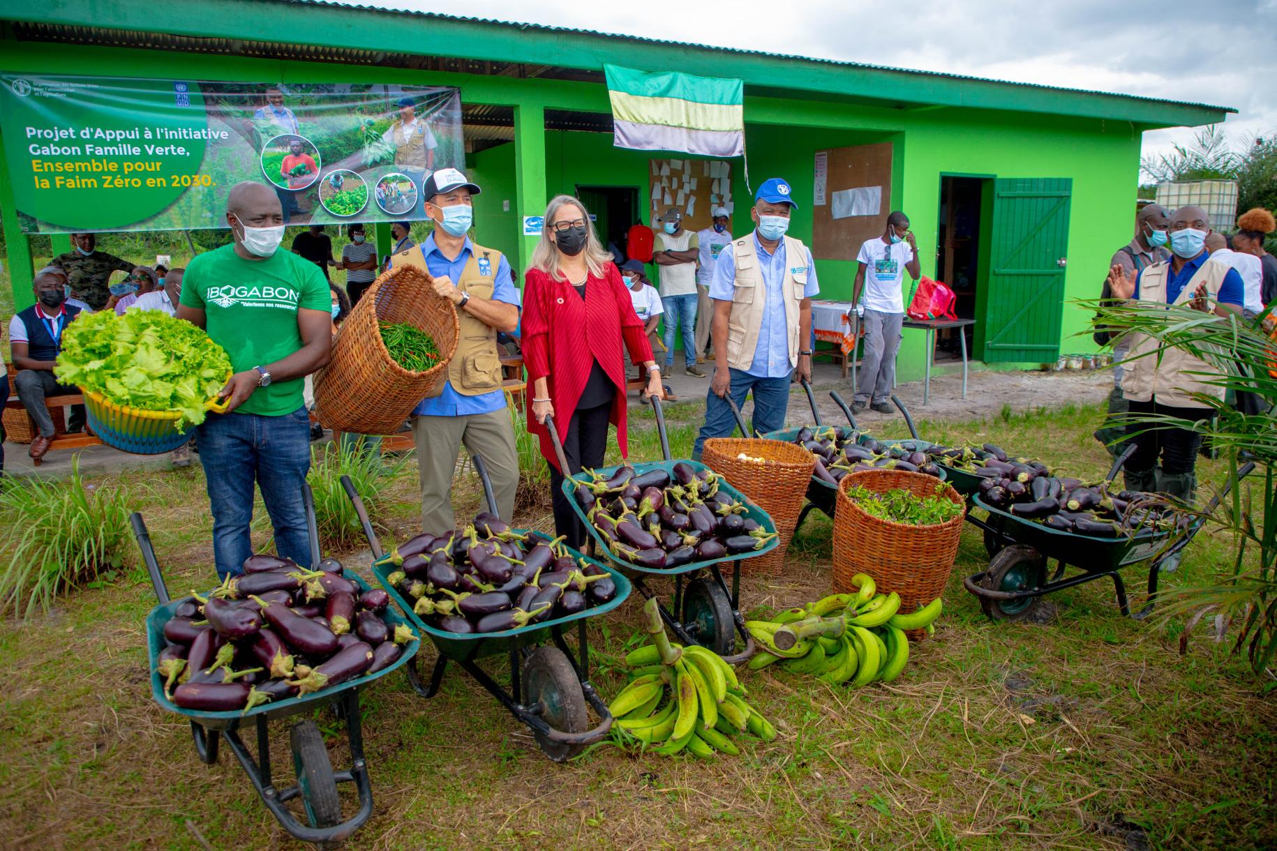 People at a small market for fruits and vegetables, in front of a green-colored building.