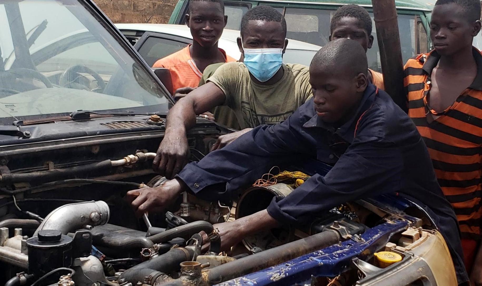 Un adolescent béninois vêtu d'une blouse de mécanien bleue et entouré de plusieurs de ses camarades est penché sur le moteur d'un véhicule et le répare.