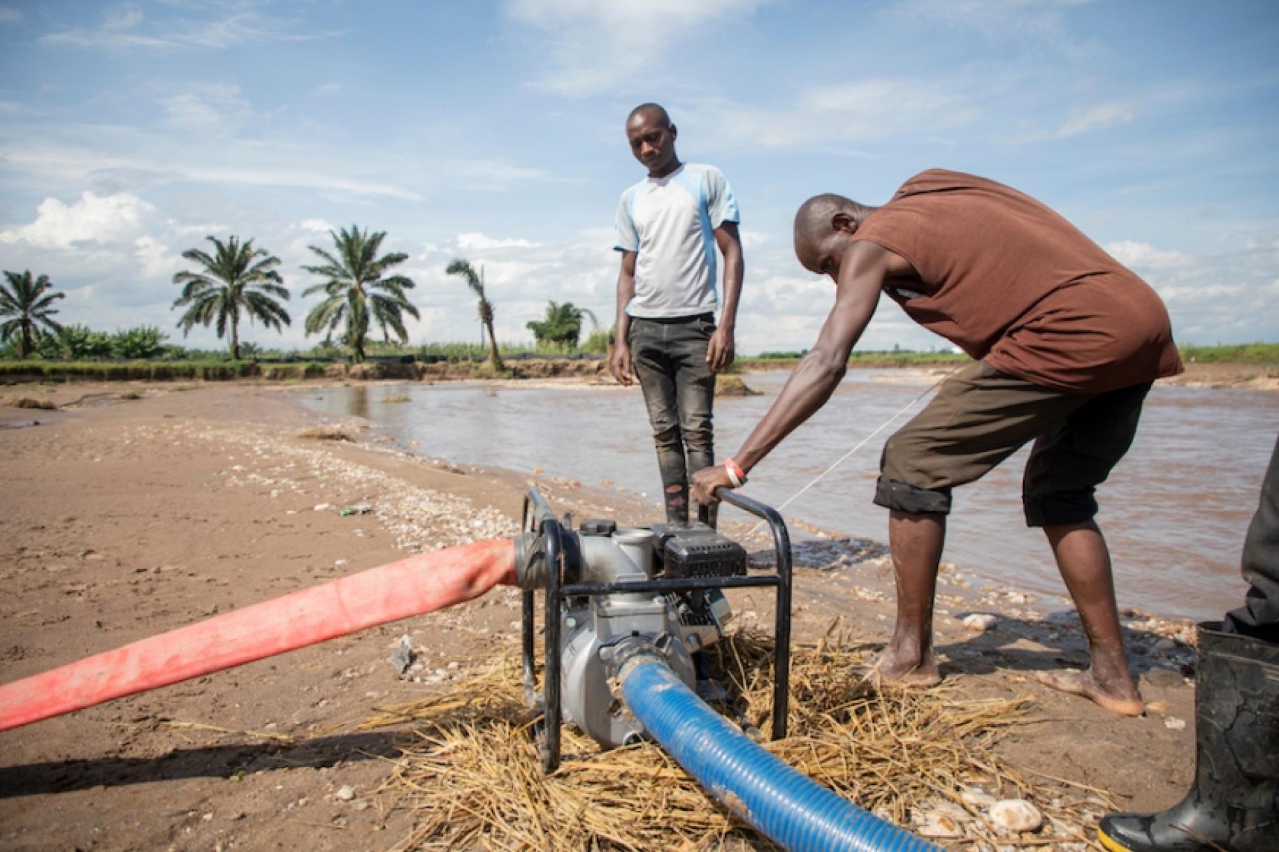 In Burundi, near a pond lined with palm trees, a man leans over the motor of a water collection system to make it work, while another man watches.