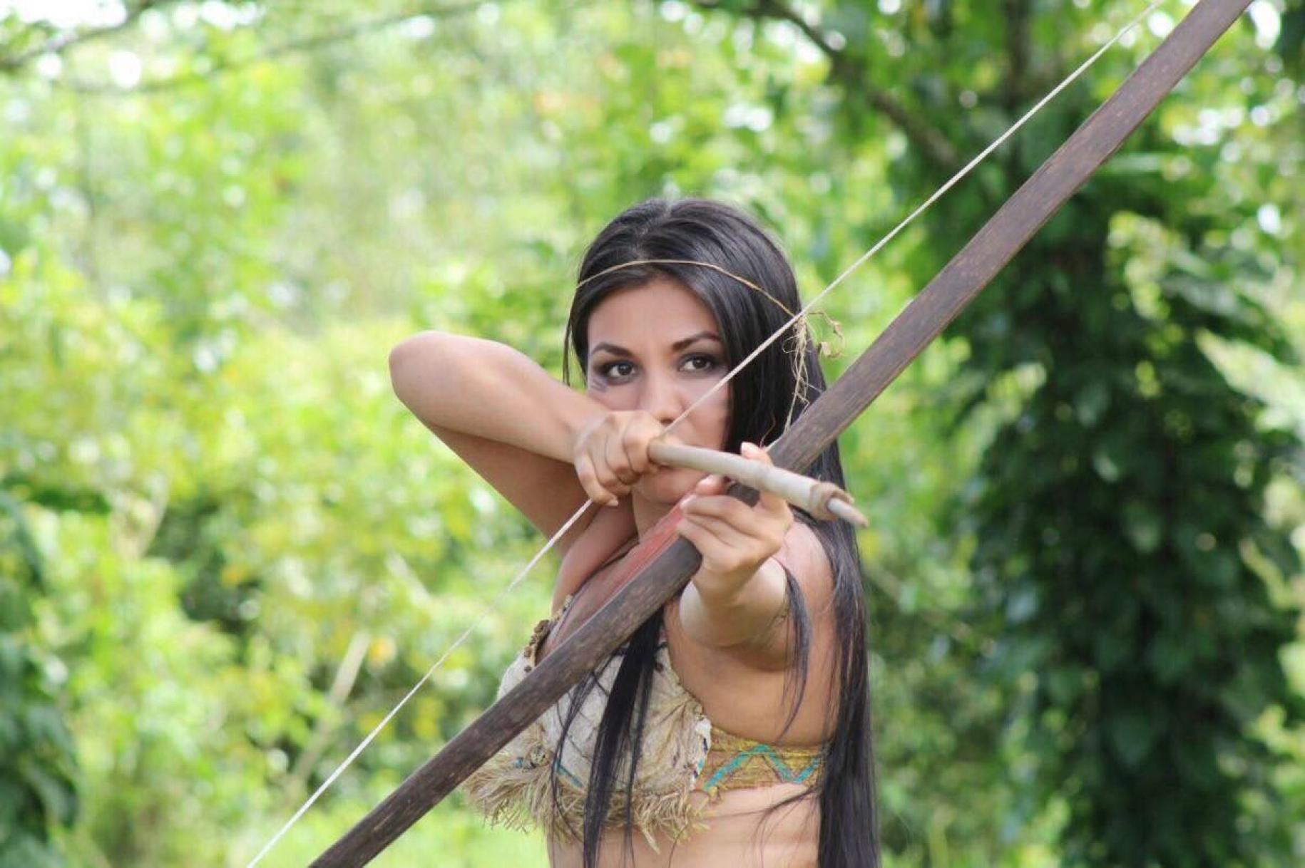 En medio de un paraje natural, una mujer enfocada en apuntar con un arco y una flecha.