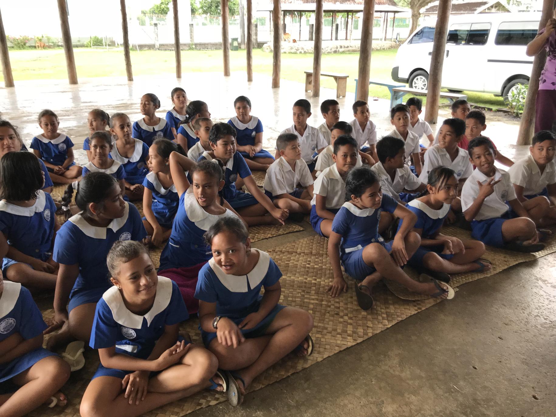 Niños y niñas con uniforme escolar sentados en colchonetas de bambú en el suelo.