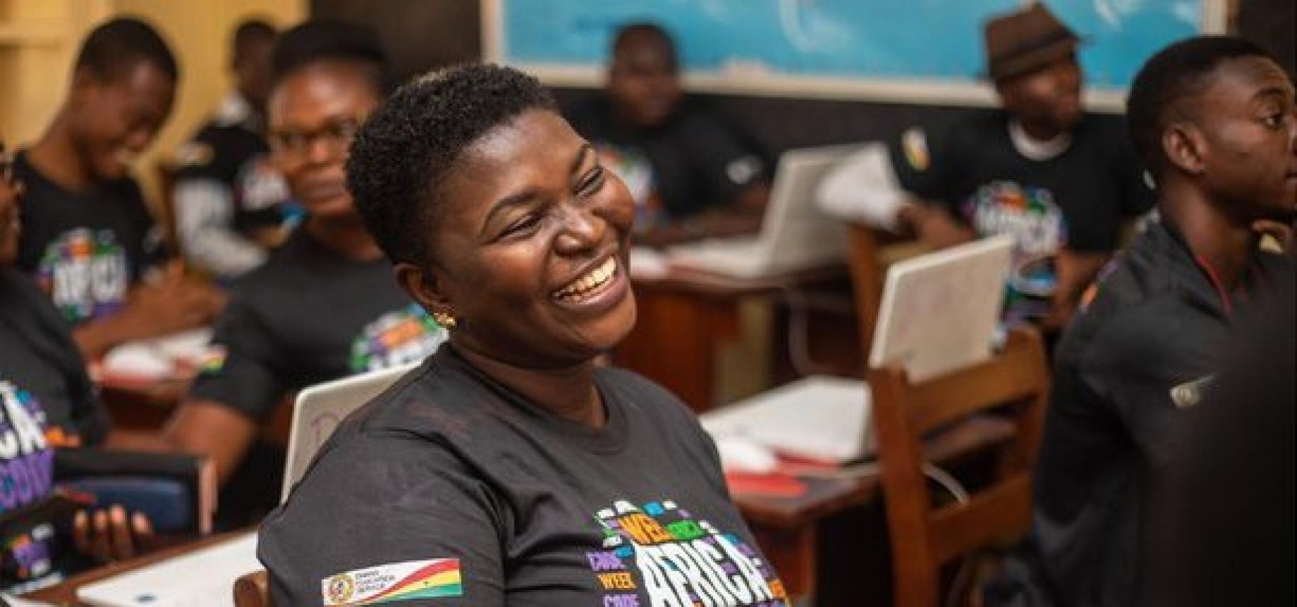 A young woman with very short hair wearing a t-shirt featuring the Africa Code Week logo is photographed in a classroom laughing.