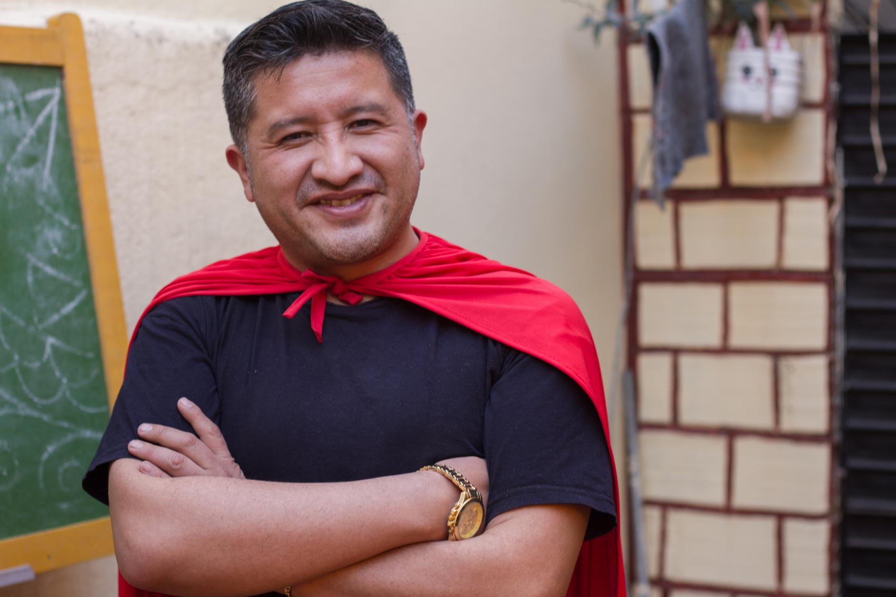 A man stands smiling in front of a blackboard, with notes made with white chalk, and wears a red superhero cape.