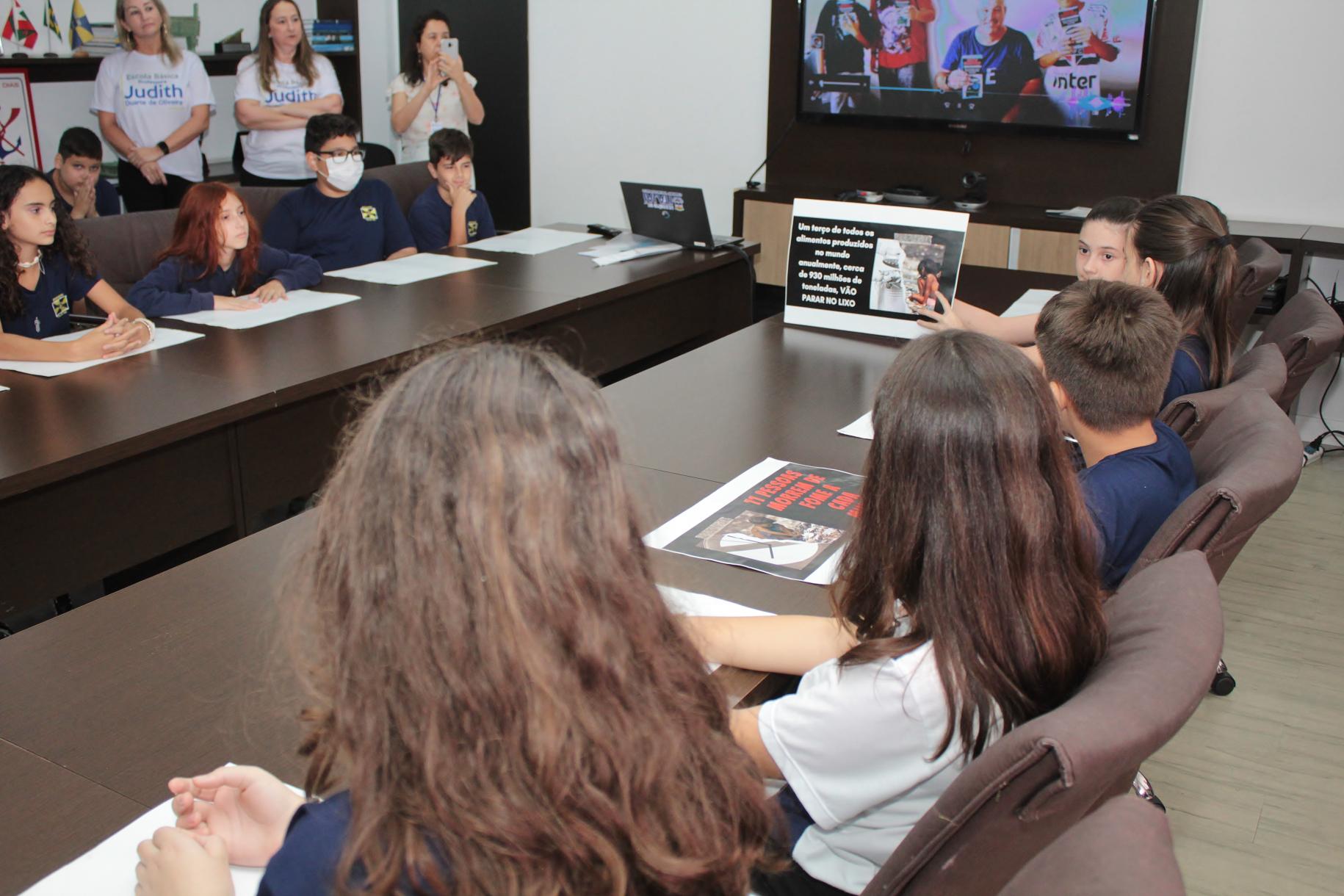 A group of children are sitting in a meeting room with their teacher at a special recognition event in the Mayor's office at  Itajaí's City Hall.
