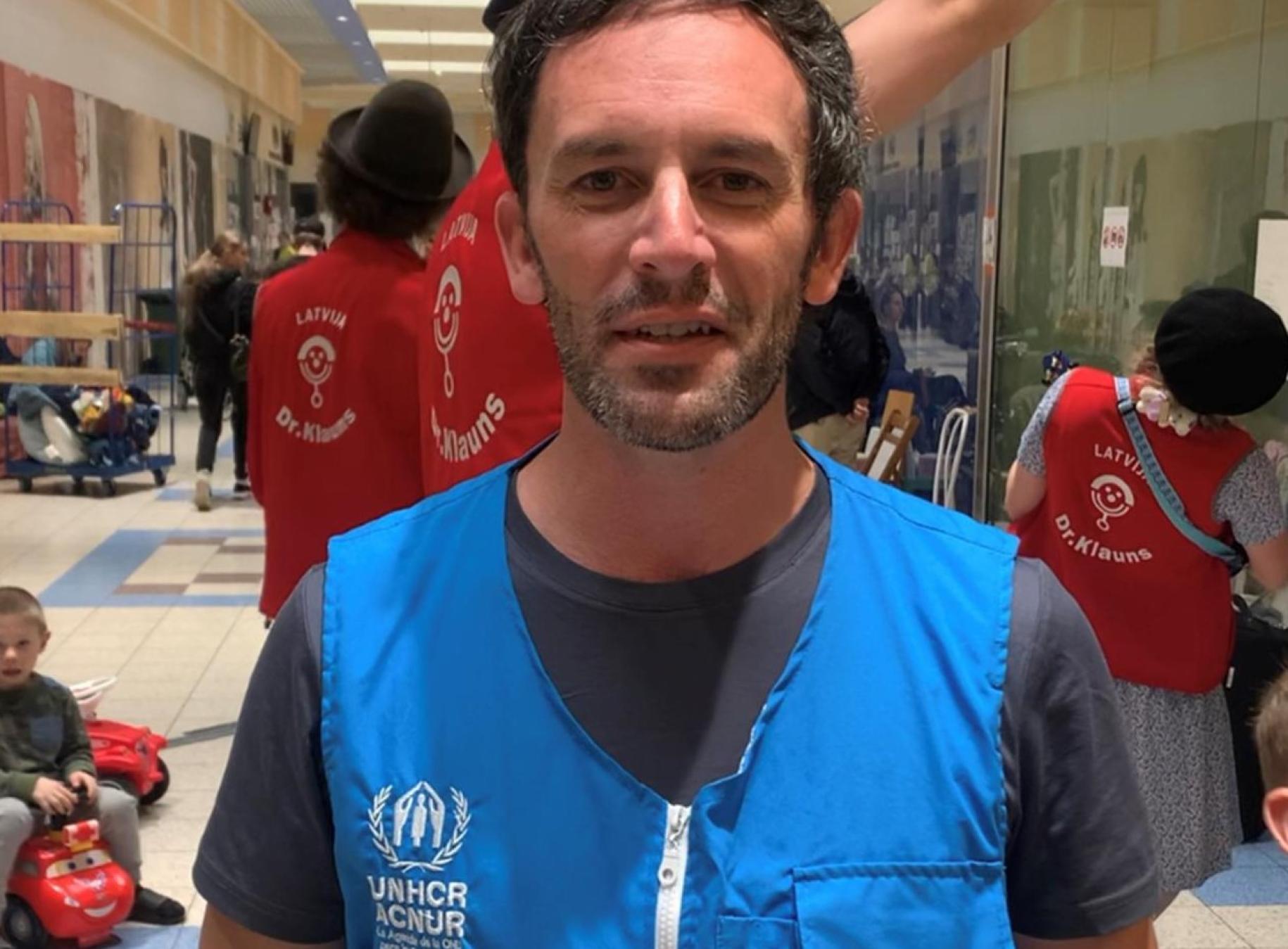 Dans un espace intérieur, un homme portant le gilet bleu du HCR s'adresse à la caméra en souriant. En arrière-plan, des personnes portant un gilet rouge s'affairent autour d'un groupe de réfugiés.