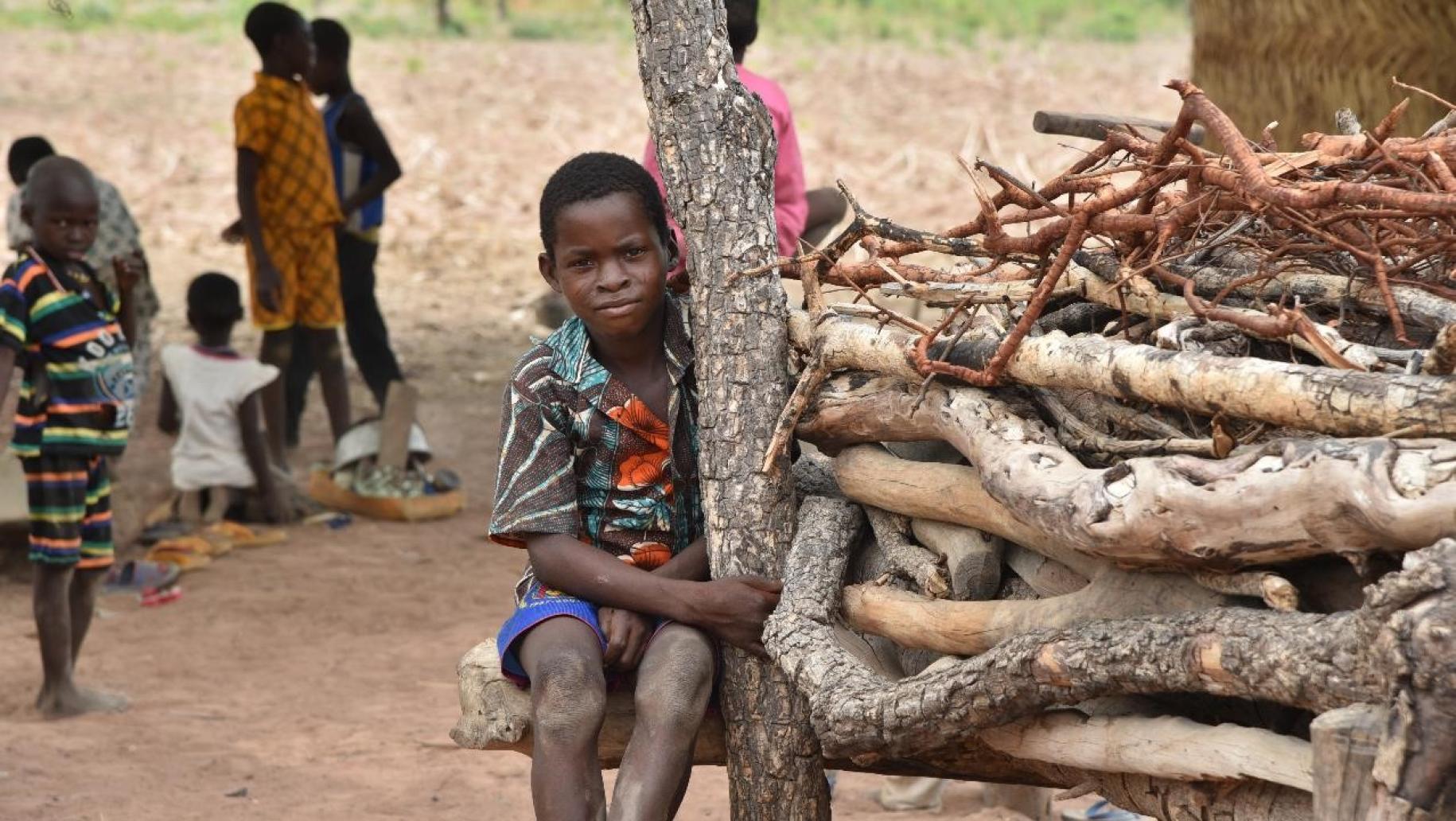 Un jeune garçon togolais est assis, face caméra, sur un amas de branches en bois à proximité d’autres enfants, en extérieur.